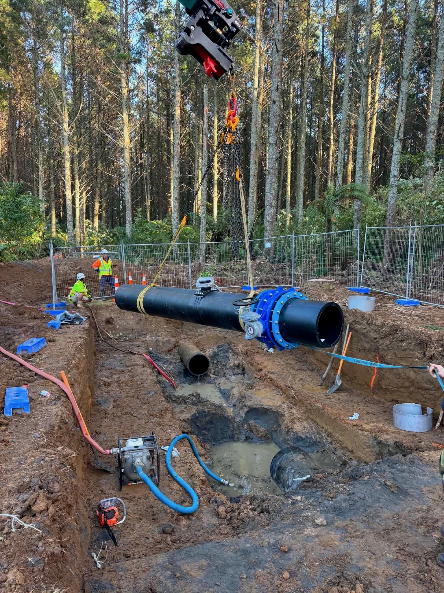 Large pipe section with valves and a blue flange suspended above an excavated trench, with workers nearby.