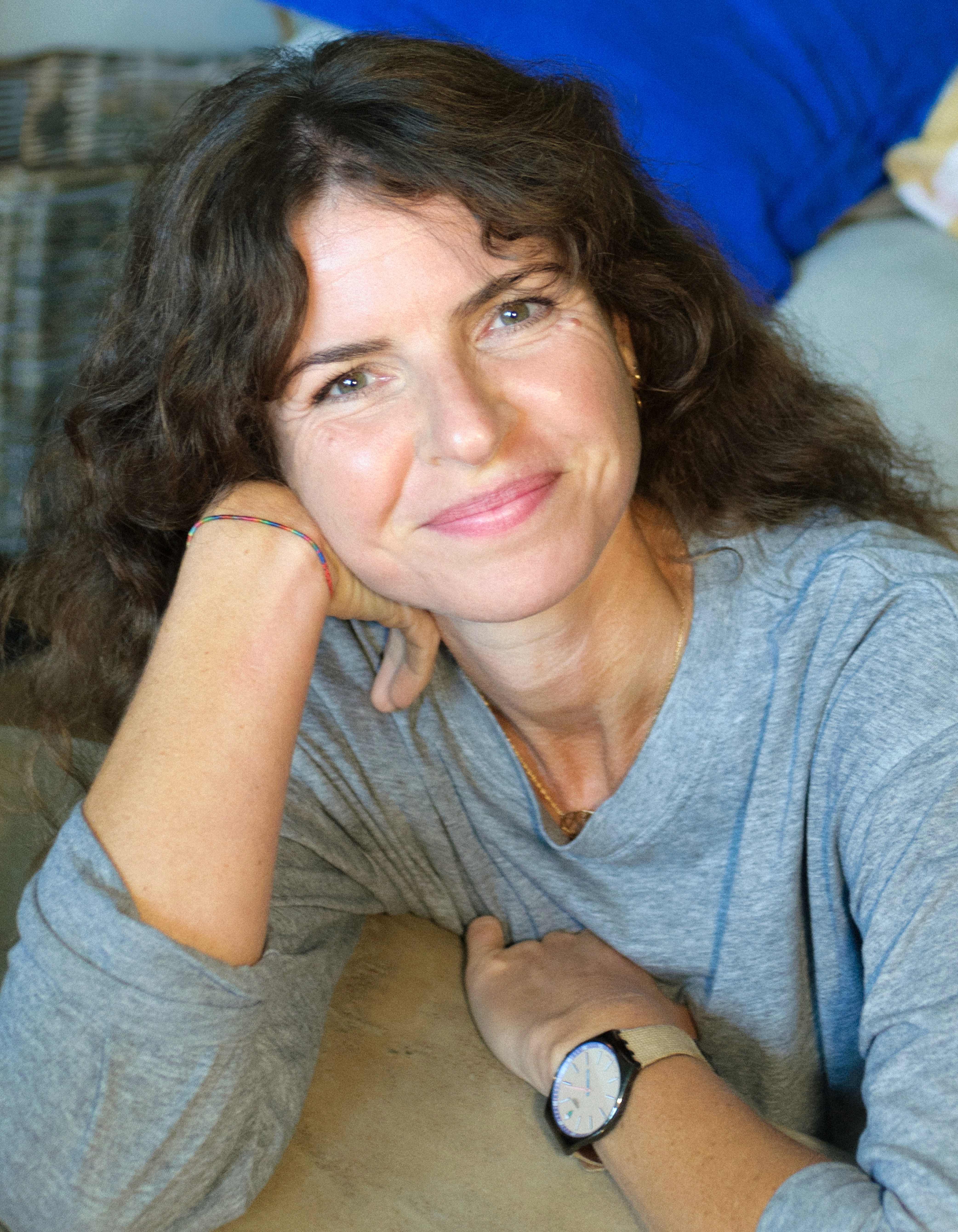 A close up colour portrait of Lucy Smith, who is sitting on the floor, with her head on her hand and her elbow resting on a low coffee table. Lucy has long mid-brown wavy hair, lightly tanned skin and blue eyes. Her expression is soft and she is smiling in a friendly and approachable way.