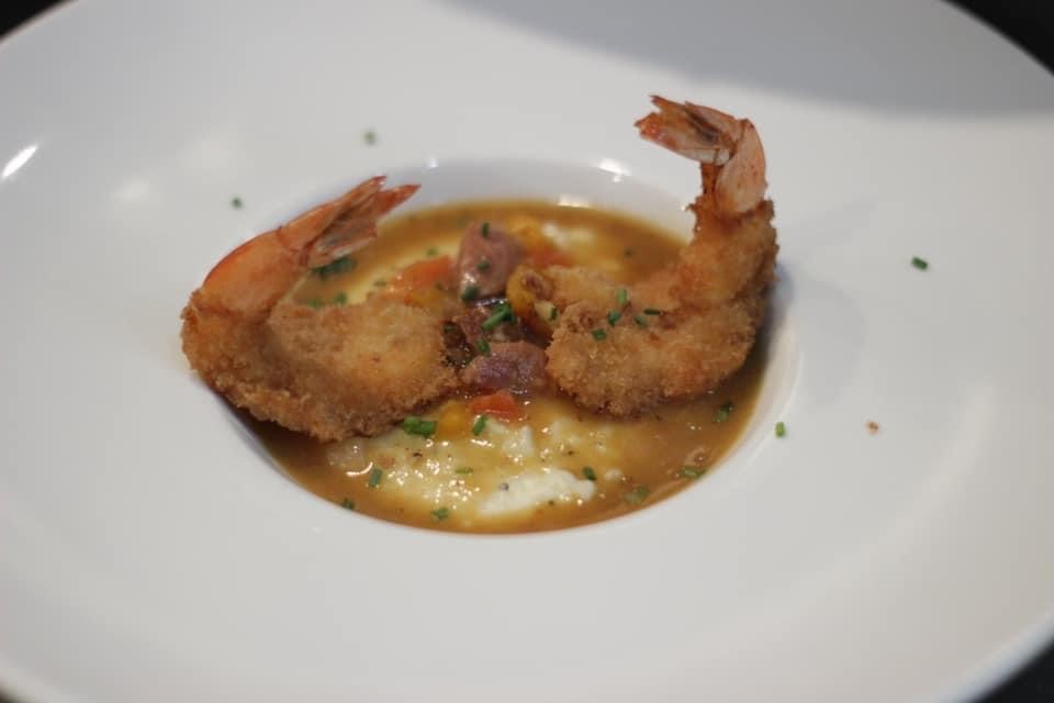 Shrimp and grits in a shallow white bowl. Fried shrimp encircle grits and gravy with sausage and tomatoes.