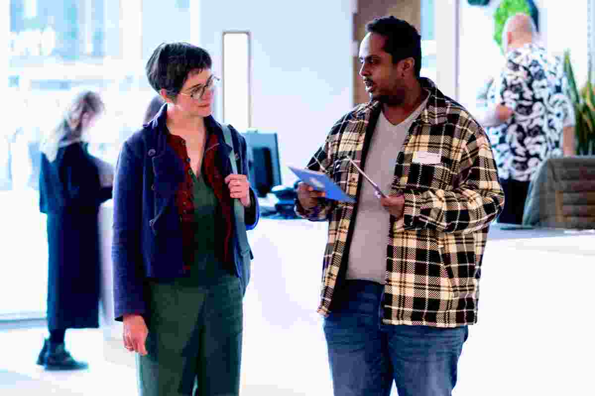 Two people standing and talking in a bright indoor event space. One person on the left is wearing a dark blue jacket over green clothing and carrying a bag over one shoulder. The person on the right is wearing a black‑and‑white check patterned jacket and jeans, holding and looking at a leaflet or programme. In the background, other attendees are visible in the open, airy venue, suggesting a casual networking or community event.