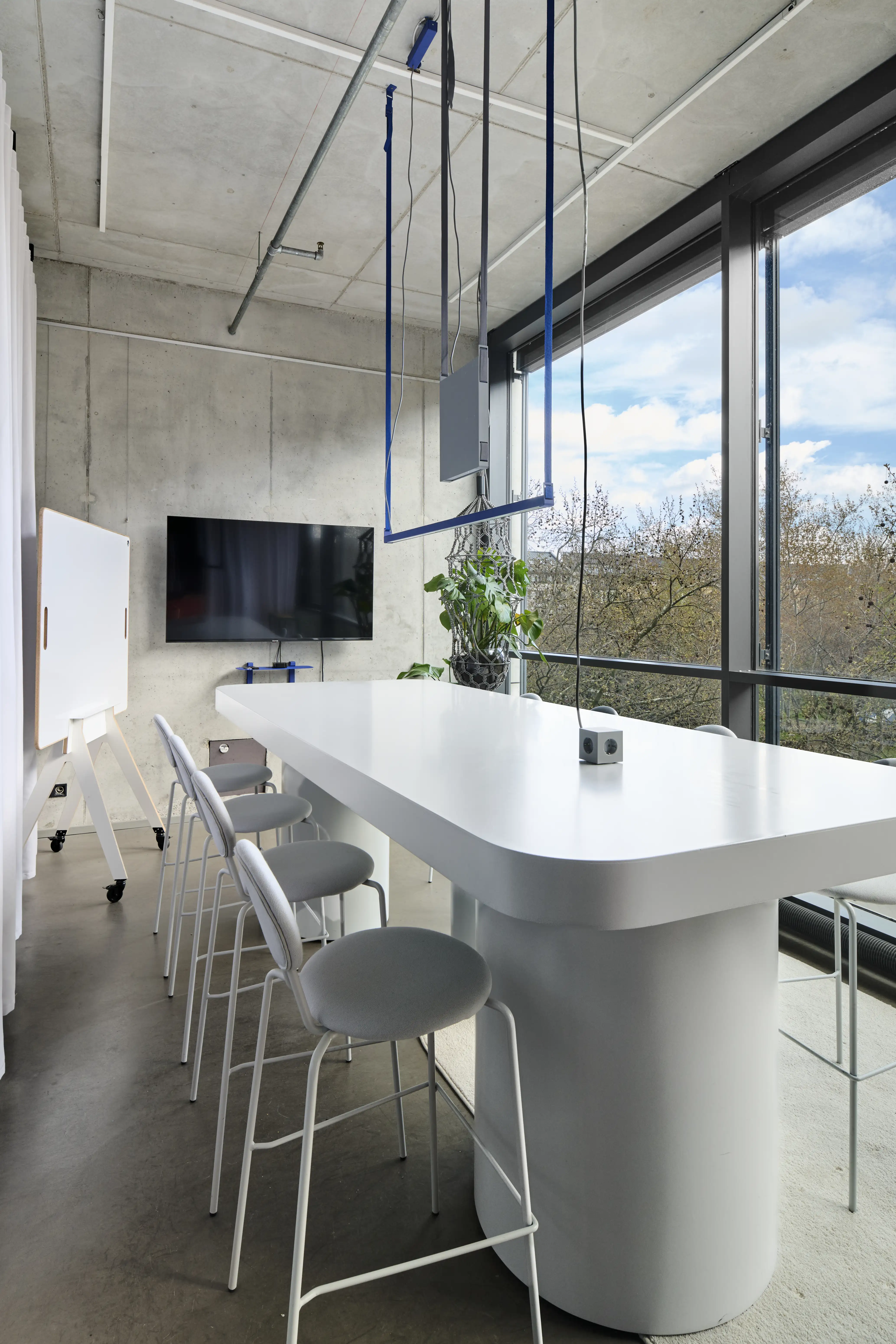 Meeting corner in the UNDPLUS SHOWROOM with a white high table and stools, wall-mounted TV and mobile whiteboard against raw concrete and floor-to-ceiling windows.