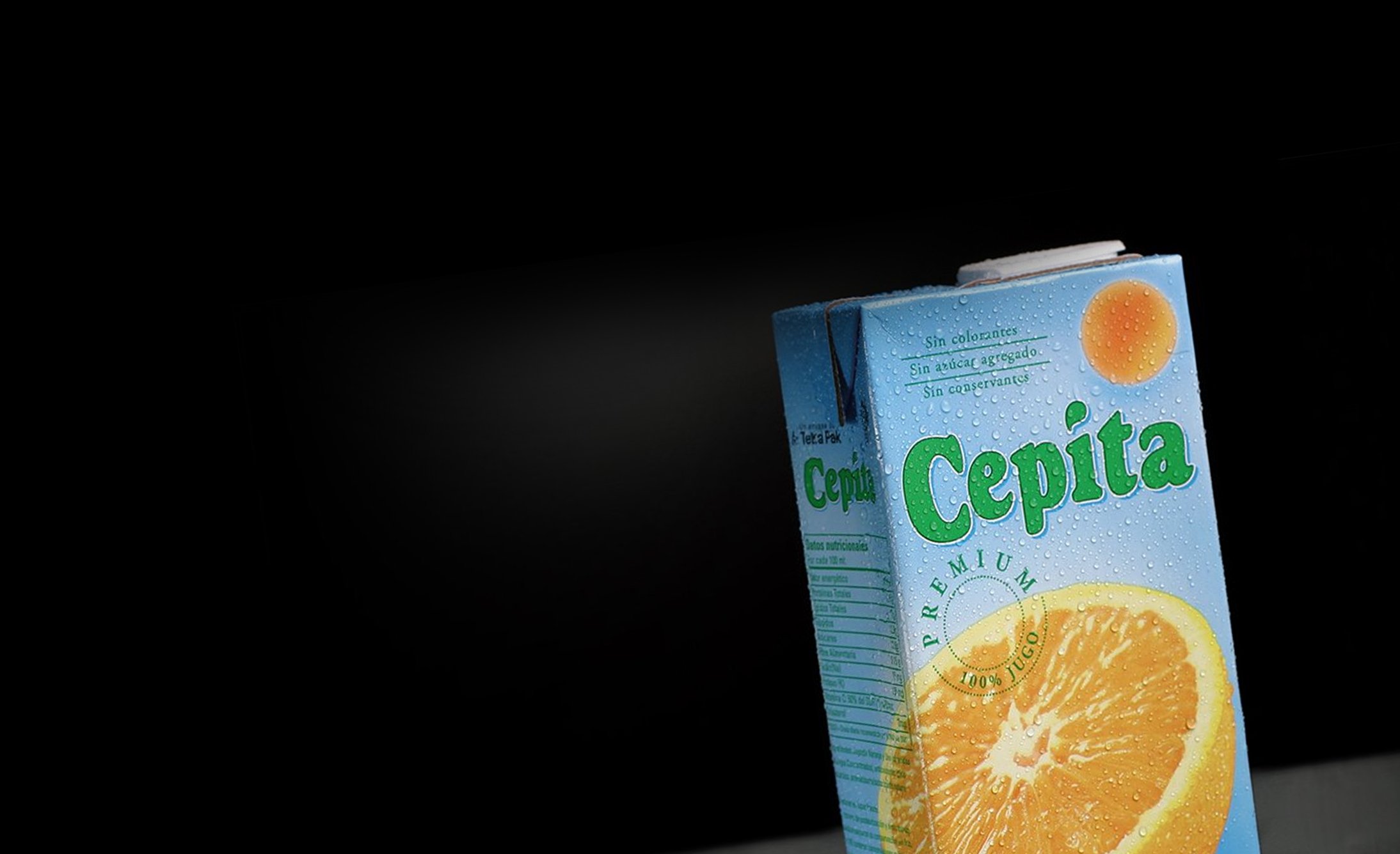 A close-up of a Cepita orange juice carton against a black background. The carton displays an image of an orange slice, with droplets suggesting freshness.