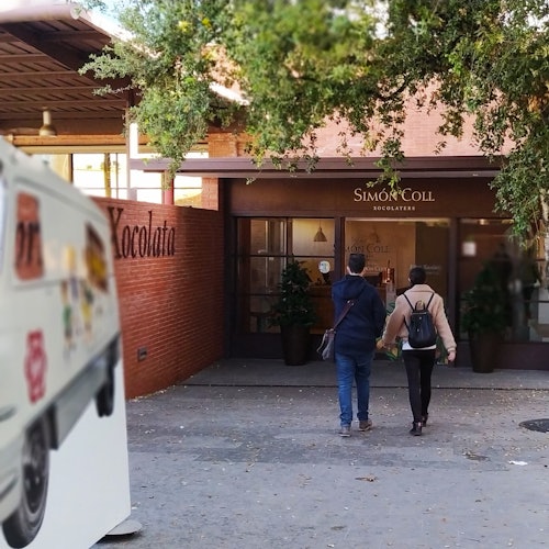 Dues persones caminant cap a l'entrada de la botiga de xocolata Simon Coll, amb un vehicle estacionat a l'esquerra i arbres al cel.
