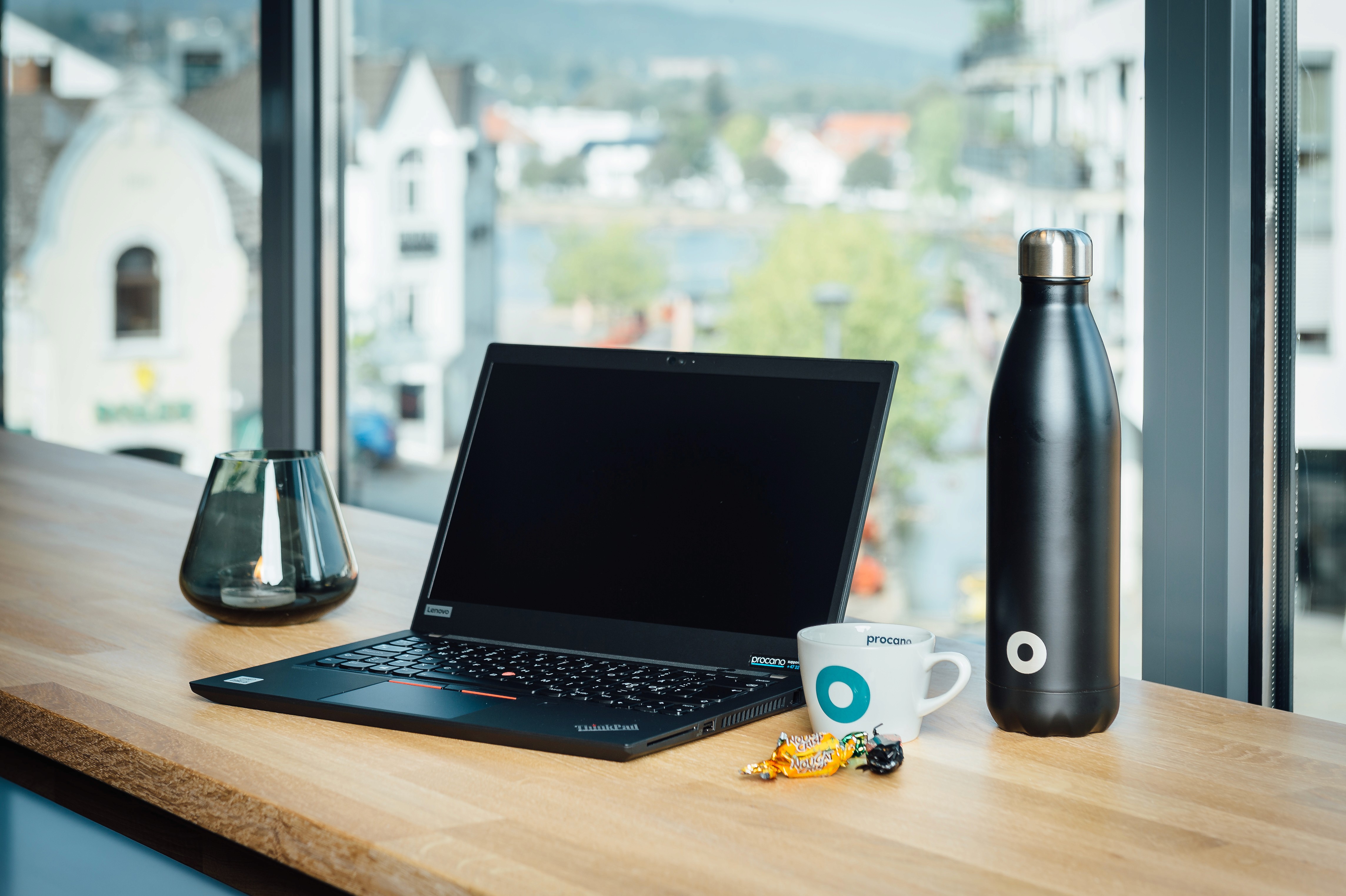 En bærbar PC, en kaffekopp med Procano‑logo, en drikkeflaske og smågodt står på en lys trebenk med utsikt mot bygninger og natur gjennom store vinduer.