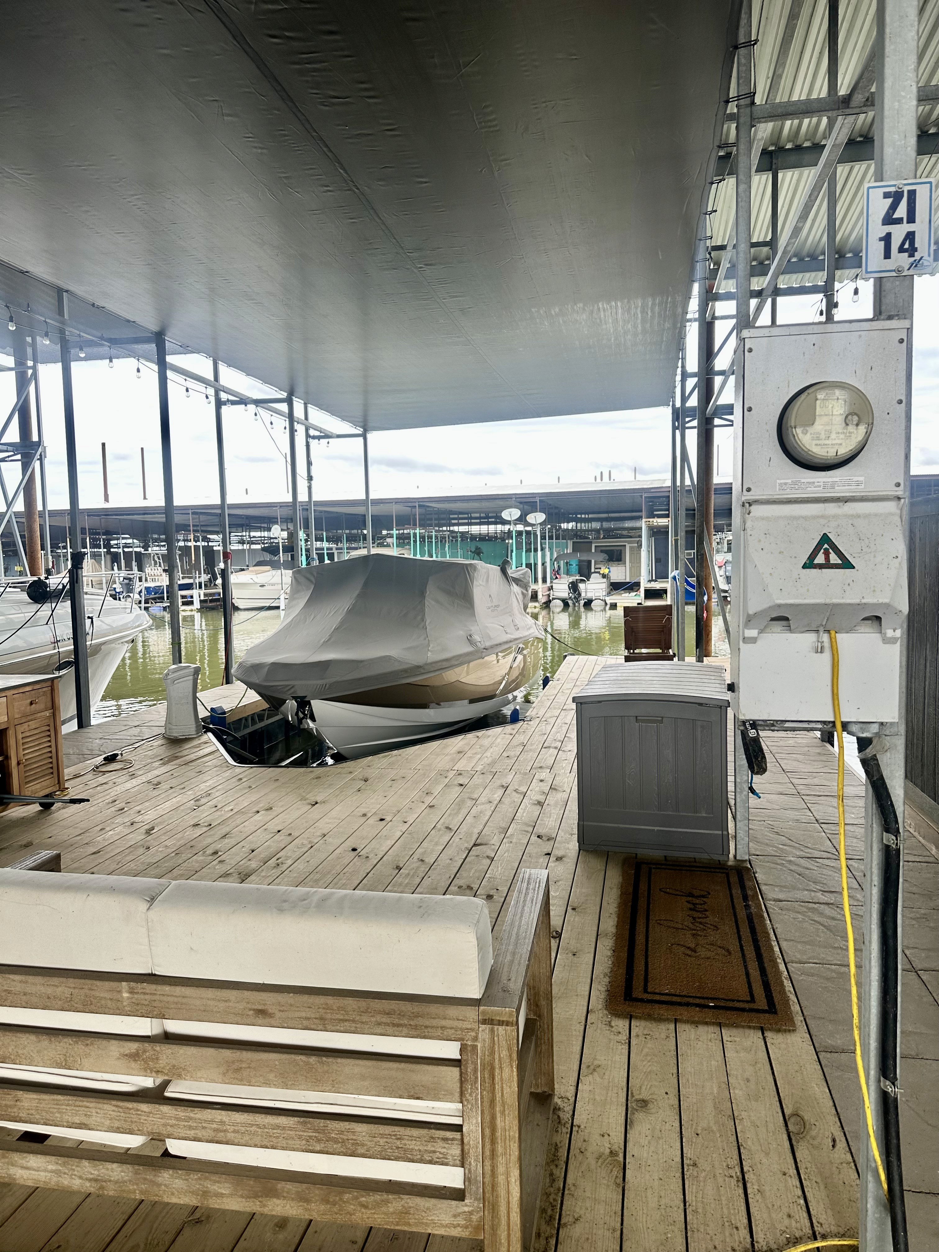 A covered marina features a wooden dock with a cushioned bench and a boat partially concealed by a tarp, surrounded by several other boats moored in the background.