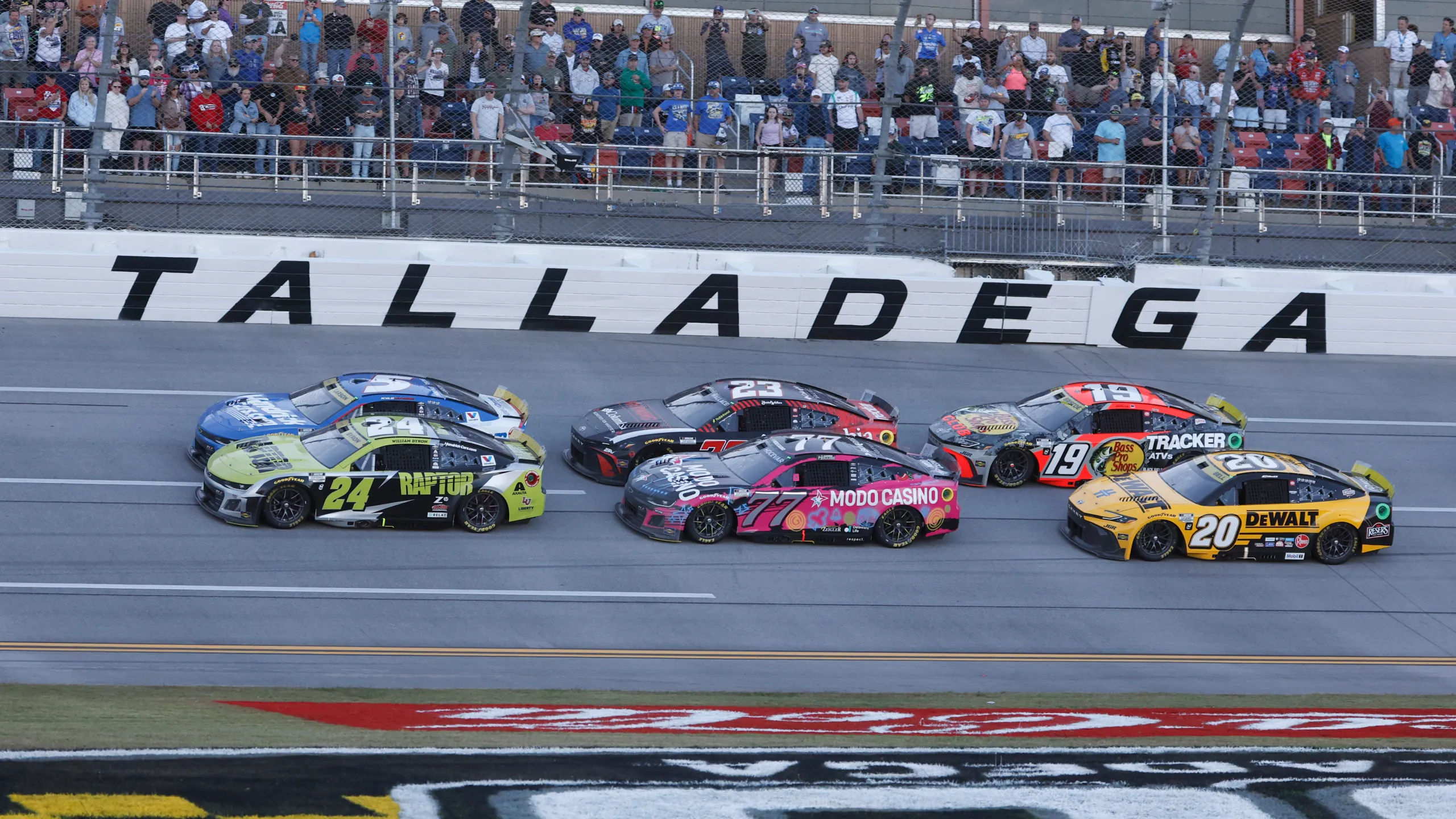 Multiple NASCAR race cars speed side by side down the Talladega Superspeedway track with fans watching from the grandstands.