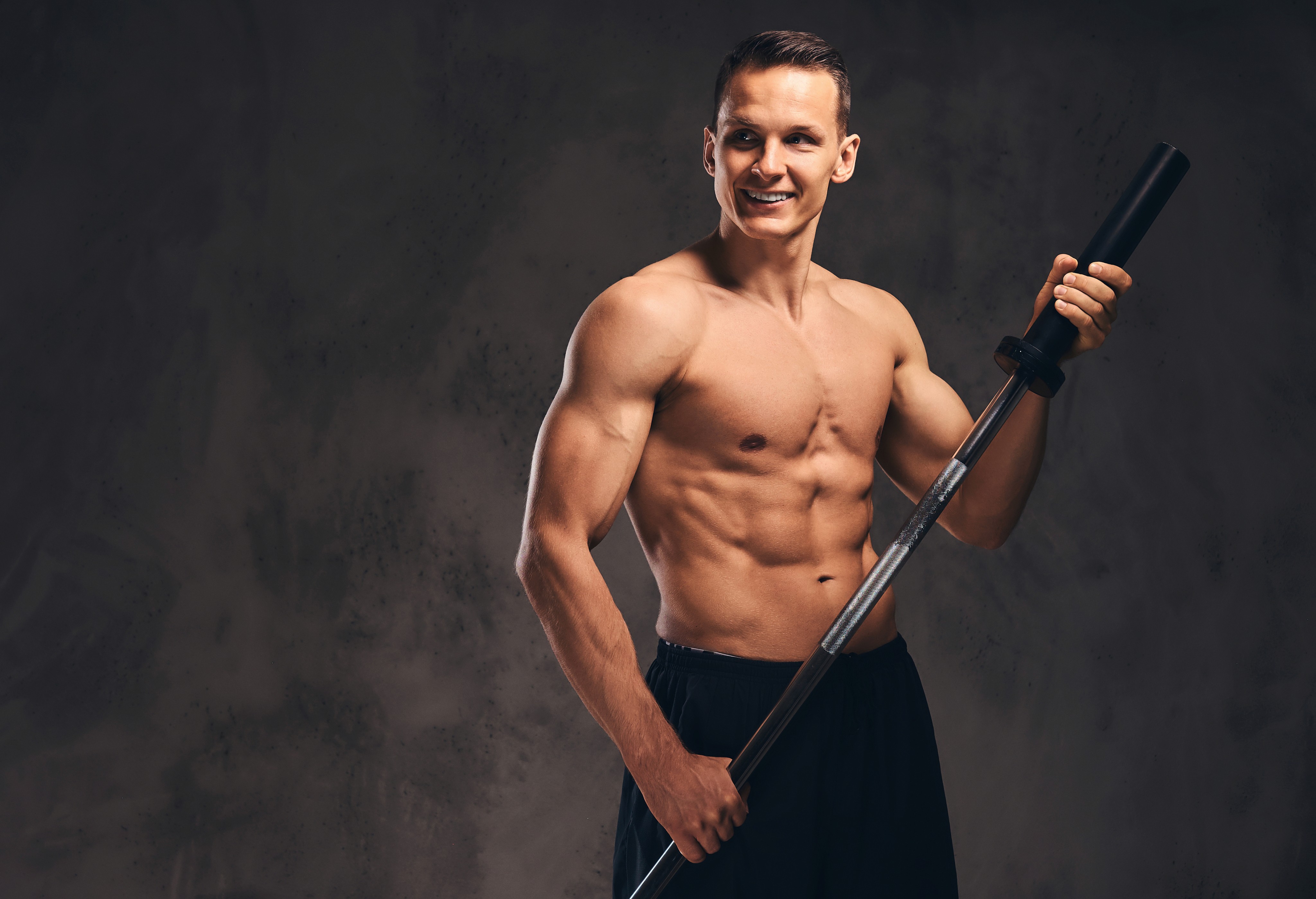 Shirtless muscular man smiling while holding a barbell against a dark background.