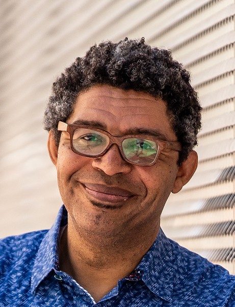 A person wearing glasses and a blue patterned shirt stands in front of a textured, striped wall, expressing a gentle smile in a well-lit environment.