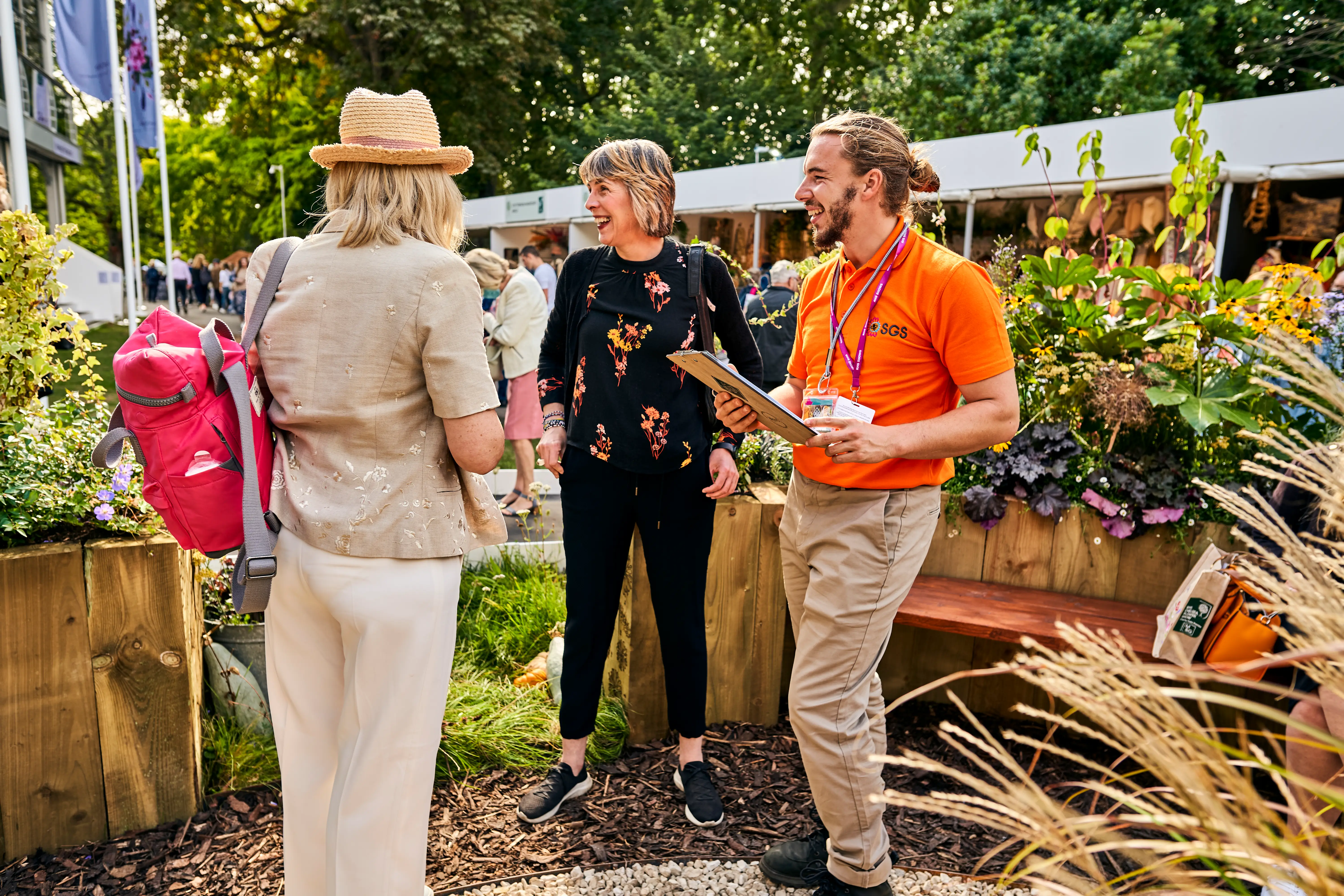 Three people converse in a garden setting, surrounded by plants and flowers, enjoying a sunny day outdoors.