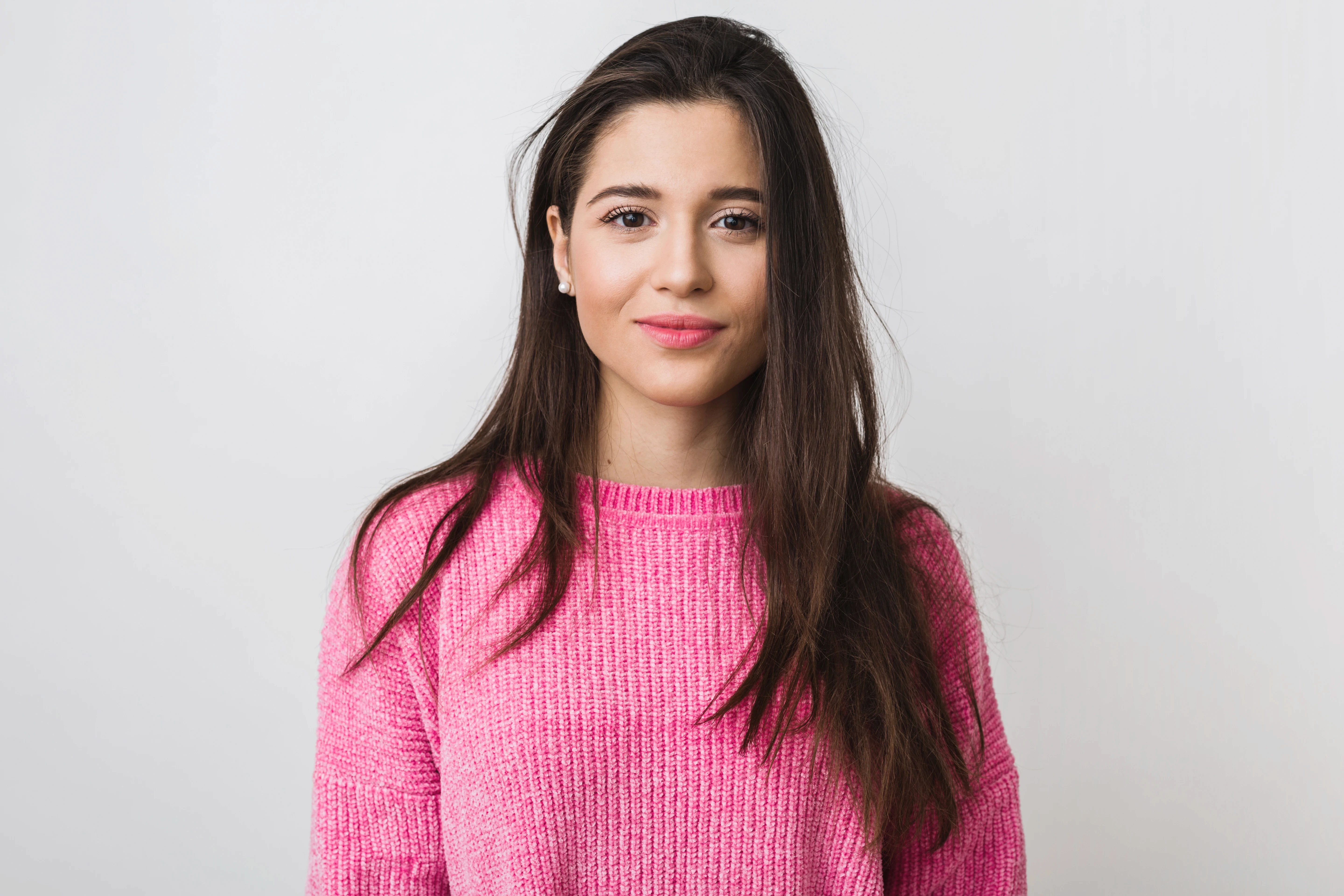 Young woman with long brown hair smiles softly, wearing a bright pink sweater. The background is plain white, creating a calm and cheerful tone.