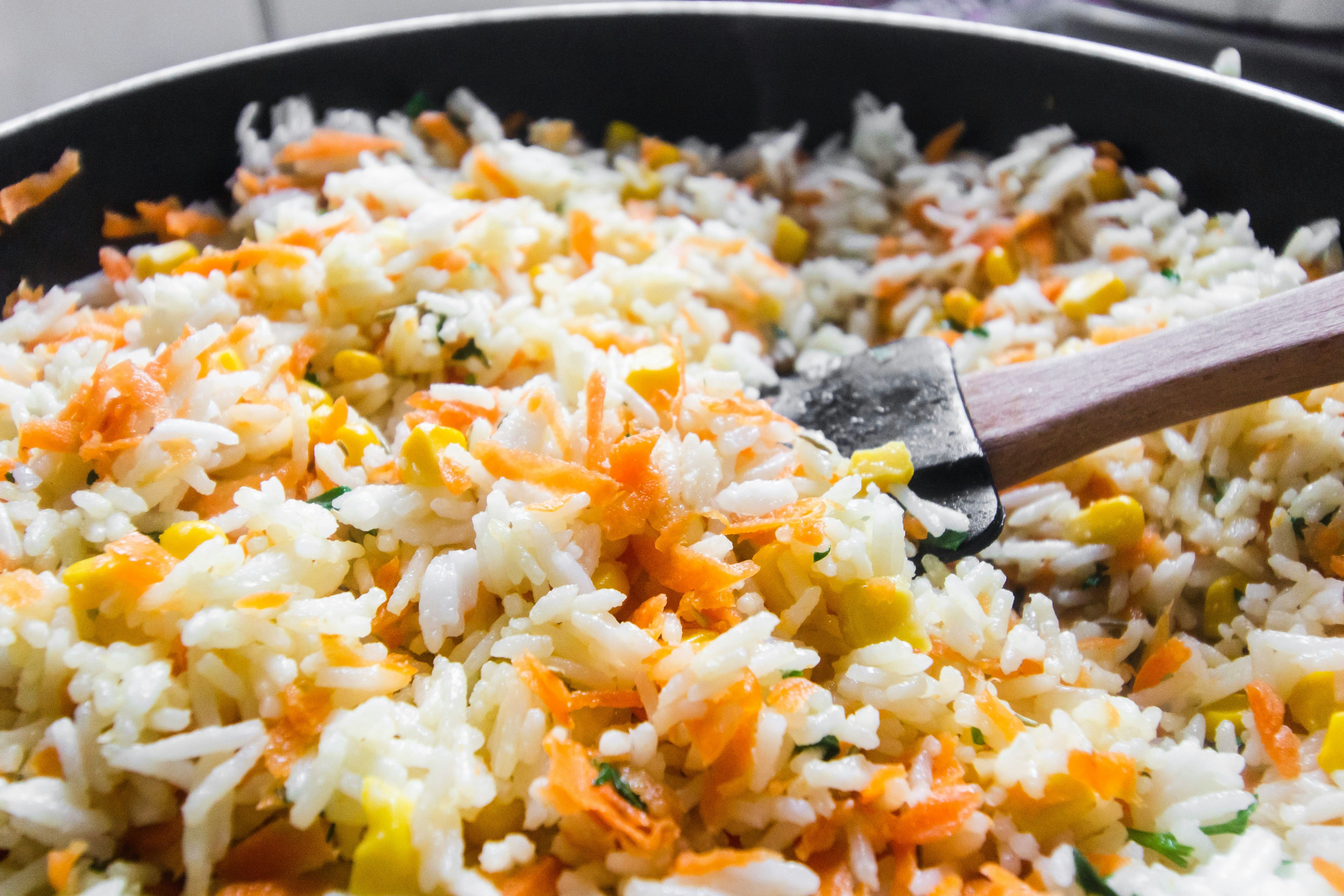 A pan filled with colorful fried rice, featuring vibrant carrots, corn, and herbs. A spatula is stirring the mixture, conveying a freshly cooked dish.
