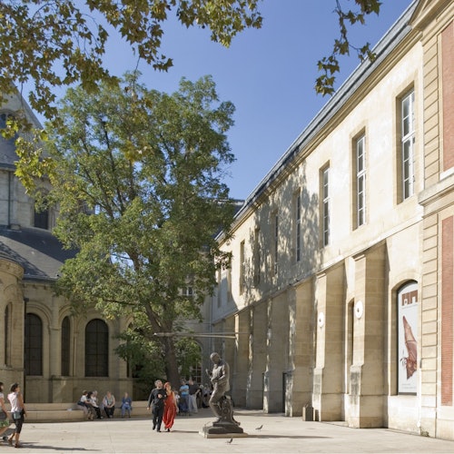 Personas caminando y sentadas en un patio soleado con árboles, una estatua y edificios históricos de piedra con ventanas arqueadas.