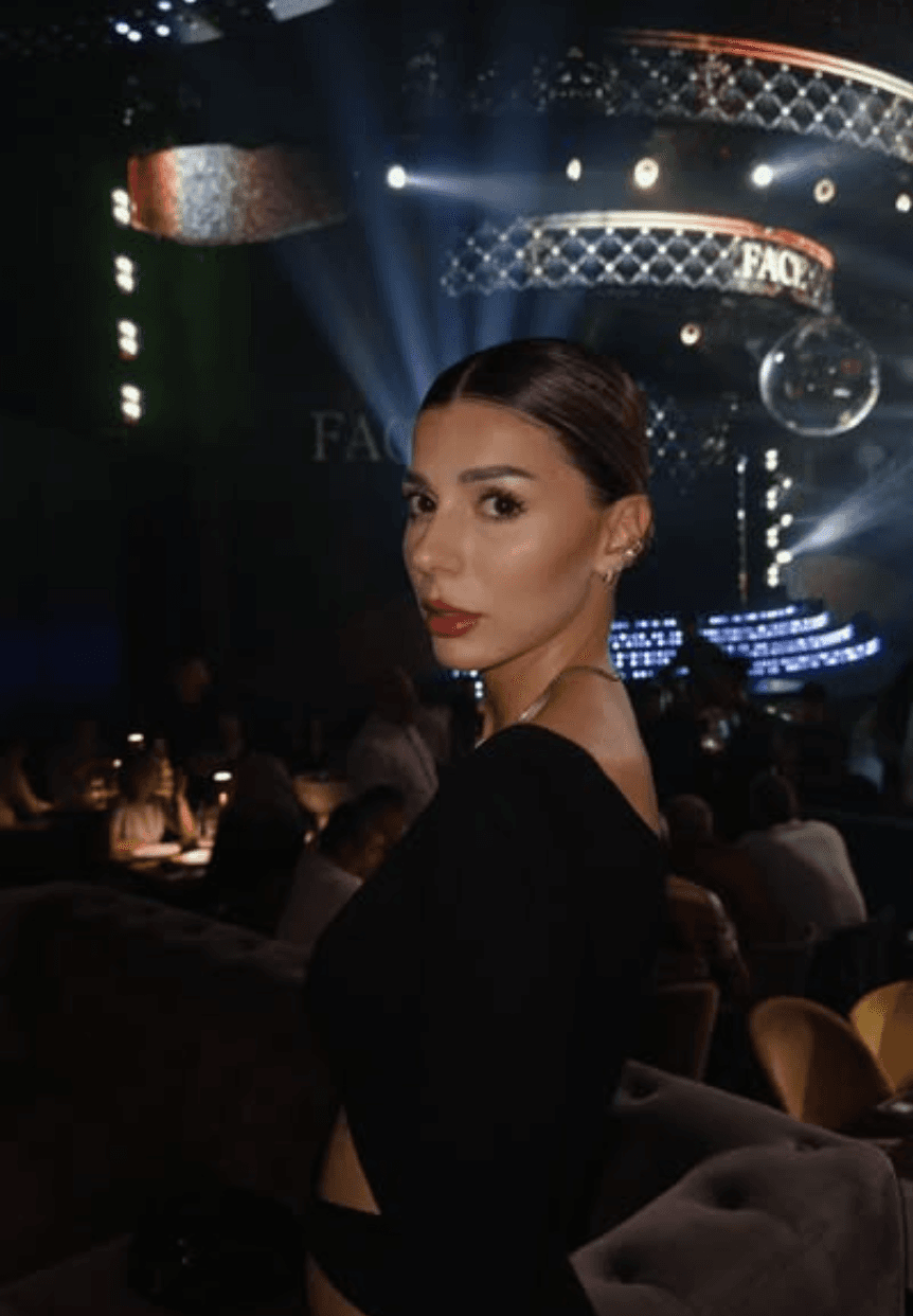 A man in a blue bomber jacket stands against a dark background. The lighting is dramatic, highlighting his focused expression. His pose is confident.