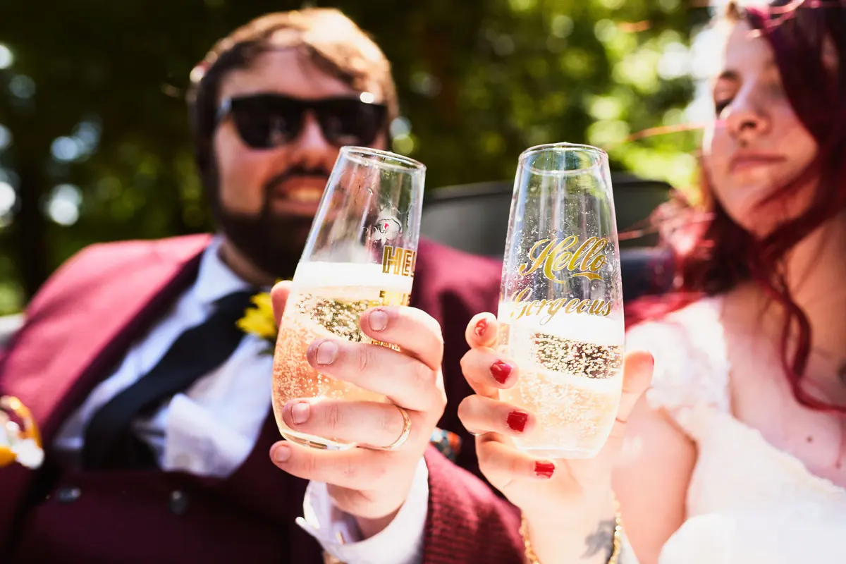 Groom in sunglasses clinking champagne glasses with the bride in the car