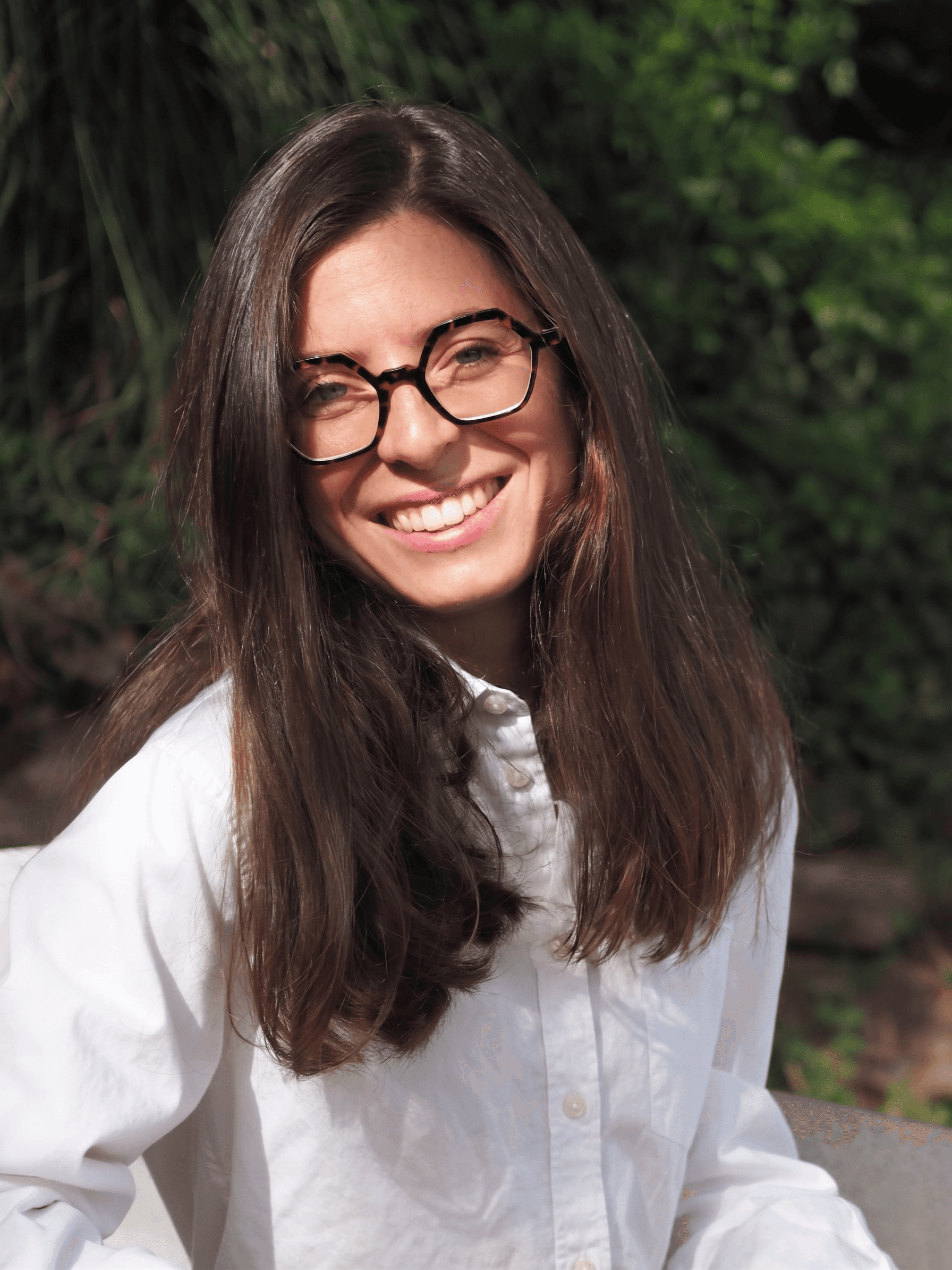 Jeune femme souriante aux longs cheveux bruns et lunettes, portant une chemise blanche, sur fond de verdure floue.
