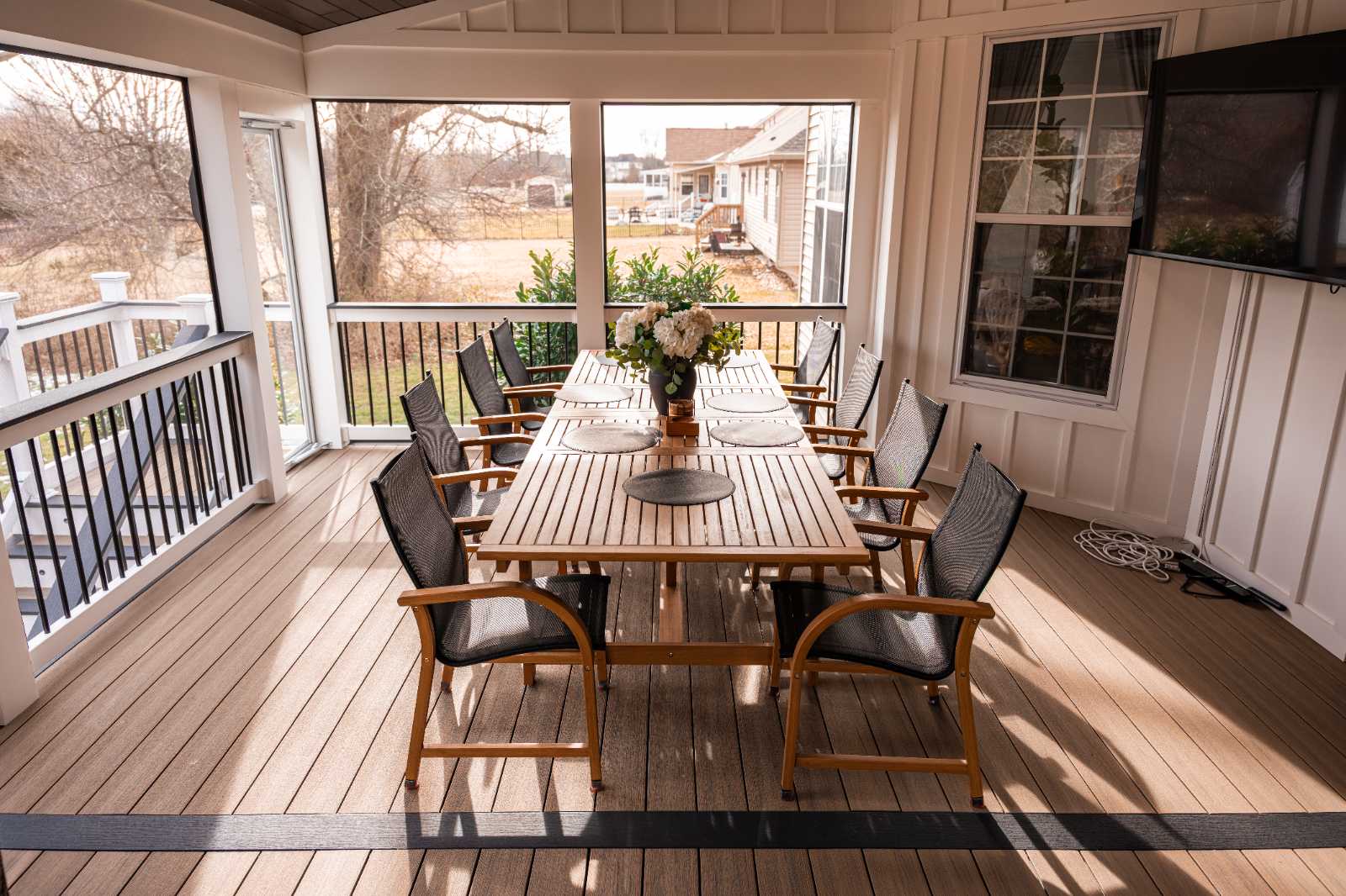 A polished wooden deck with gleaming rain-soaked planks is bordered by a white railing featuring vertical black metal balusters, set against a backdrop of lush green trees and a manicured lawn.