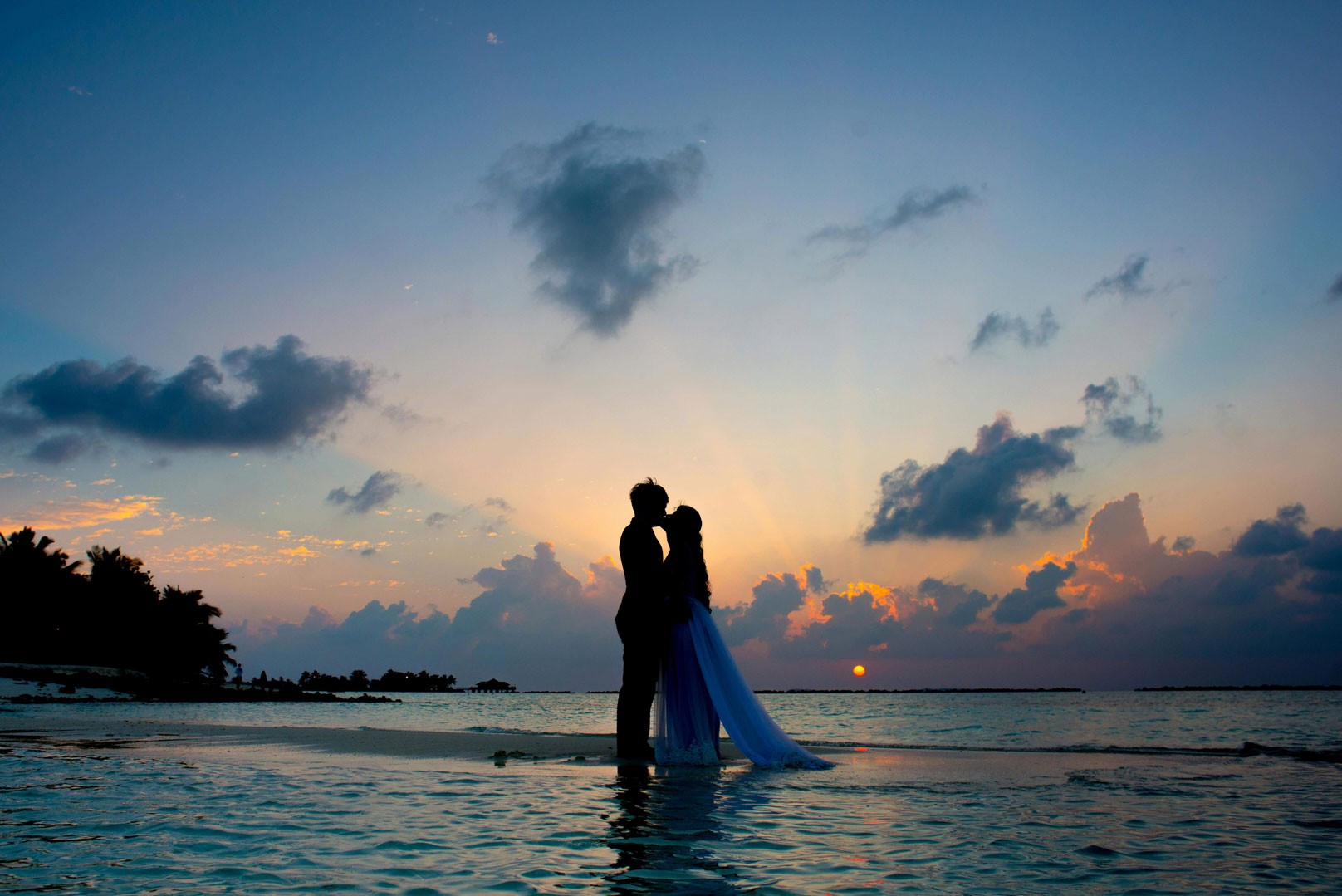 Romantic silhouette of a wedding couple kissing on a Fiji beachfront at sunset, the bride's dress flowing in the water.