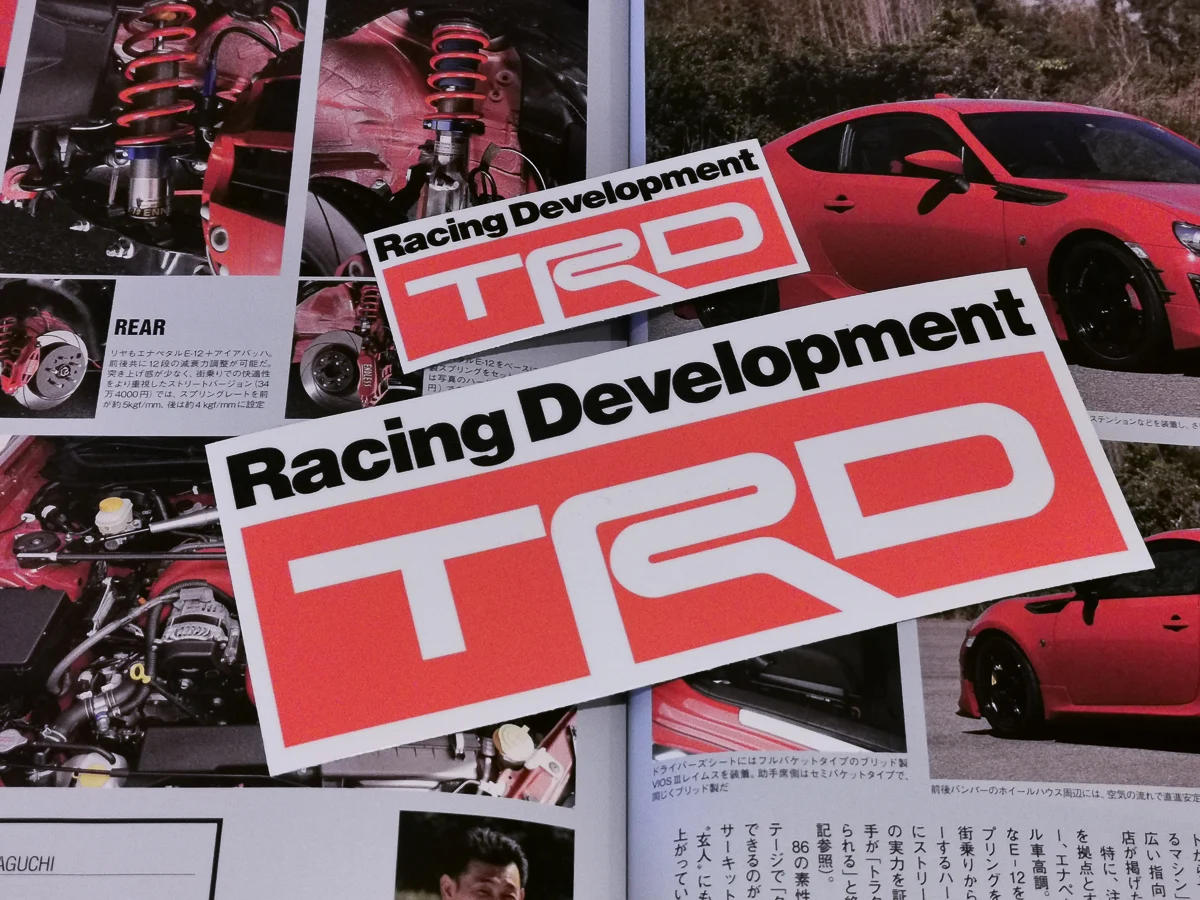 Two TRD (Toyota Racing Development) stickers placed over a magazine featuring car suspension, engine components, and a red Toyota sports car. Emphasizes TRD’s connection to performance tuning and custom parts.