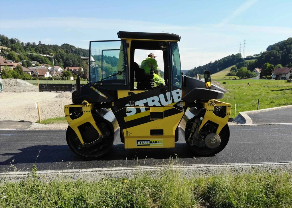 Strub Bau AG worker operating a double drum roller compactor during road surfacing in a rural Swiss landscape – precision in asphalt compaction.