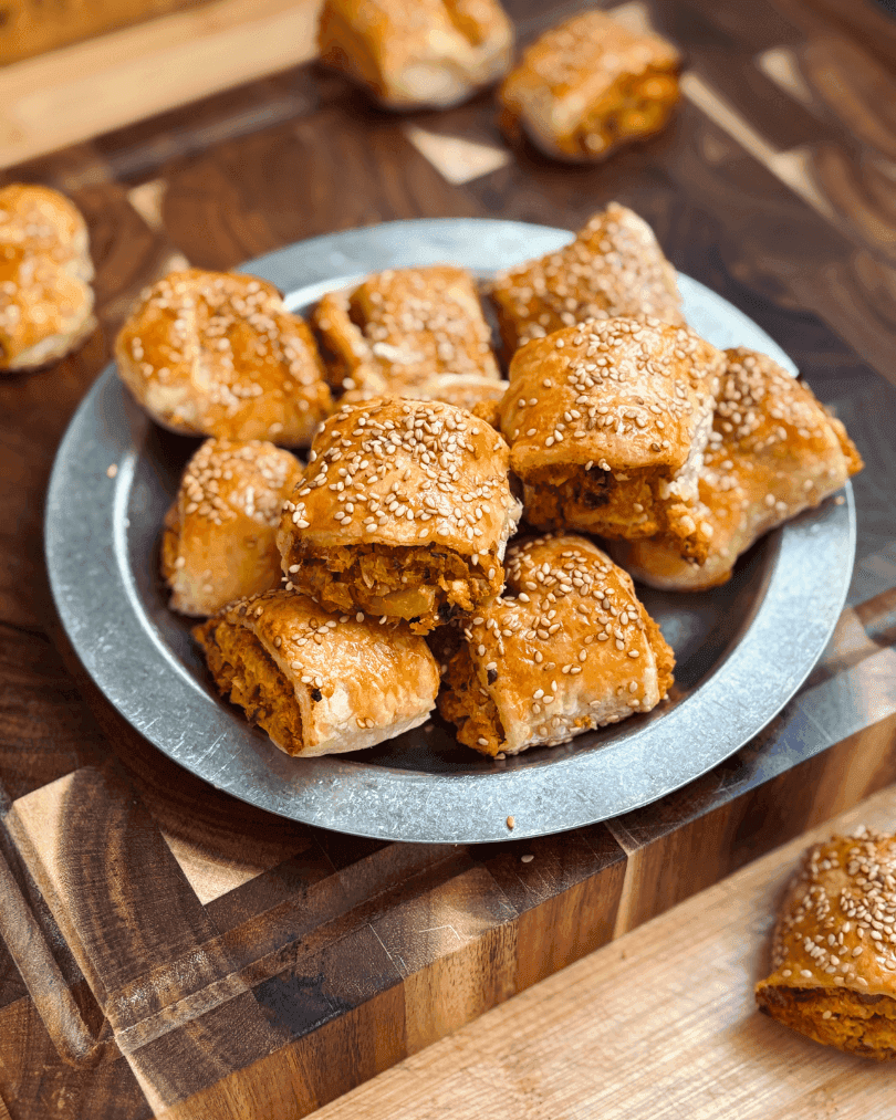 Bite-sized sweet potato and chickpea rolls baked in puff pastry, topped with sesame seeds and served on a metal plate.