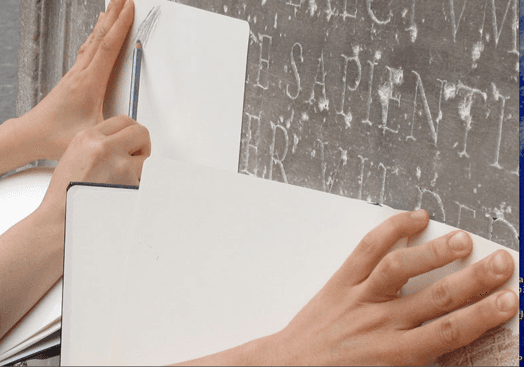 A photo of a pair of hands holding pieces of paper against an engraved wall and copying the writings with a pencil.