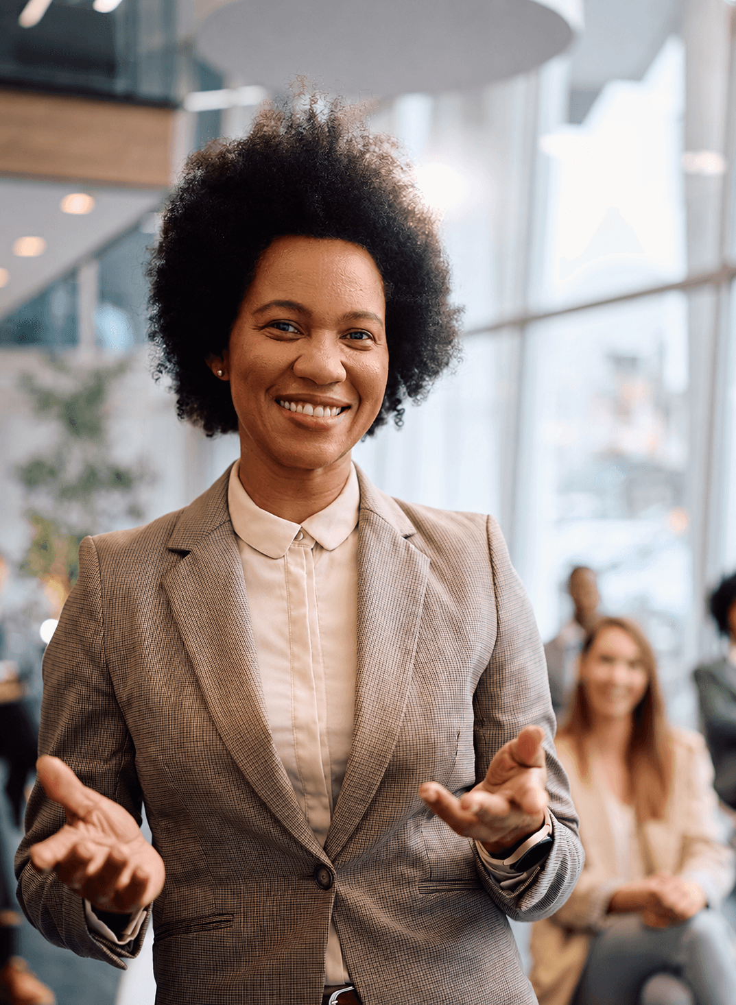 “Smiling woman with curly hair, wearing a beige plaid blazer, speaking with open hands during a presentation to a seated audience in a modern office.”