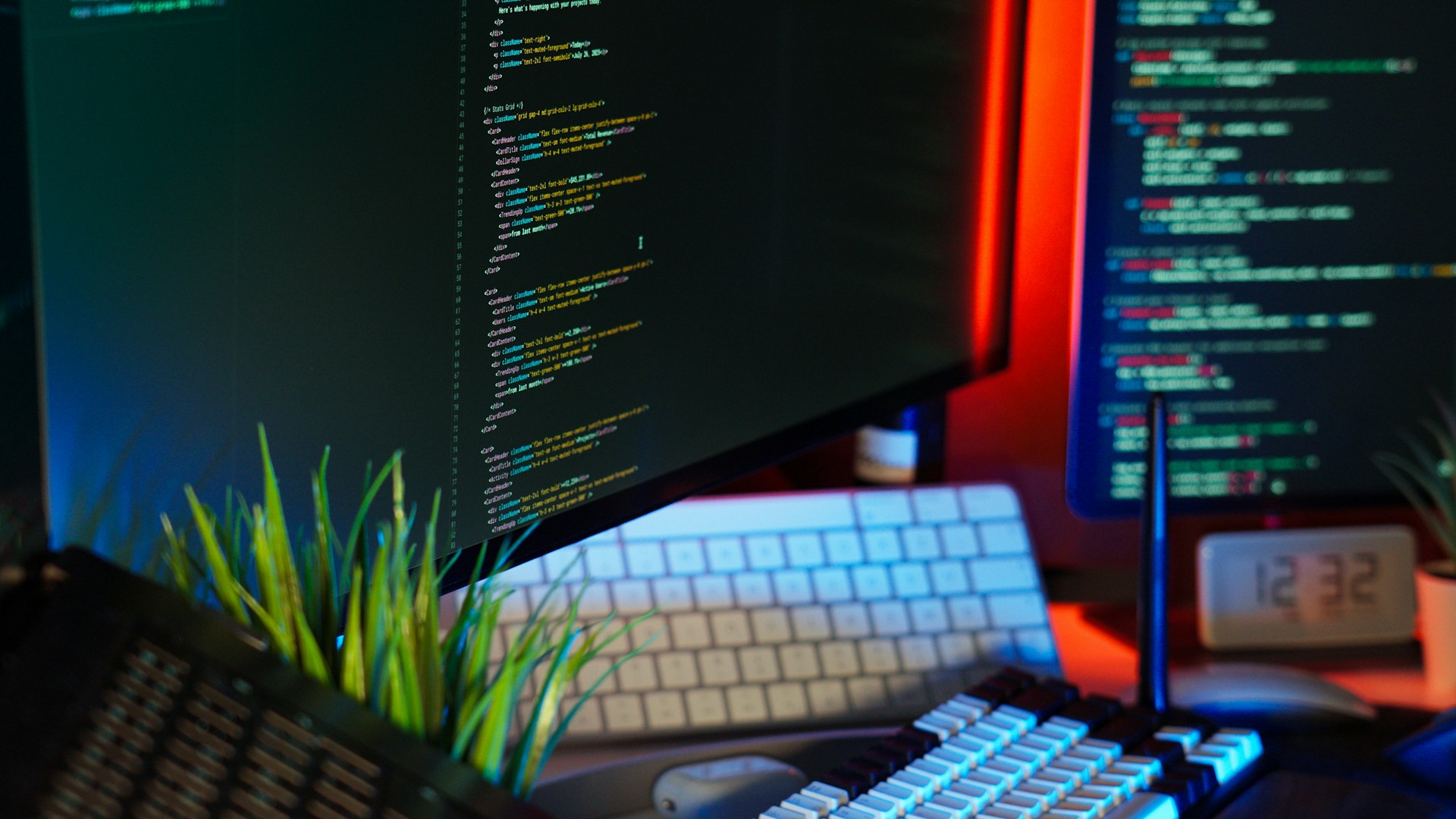A dark, close-up shot of two computer monitors displaying brightly colored code, with two keyboards and a small green plant visible on the desk, creating a programmer's workspace aesthetic.