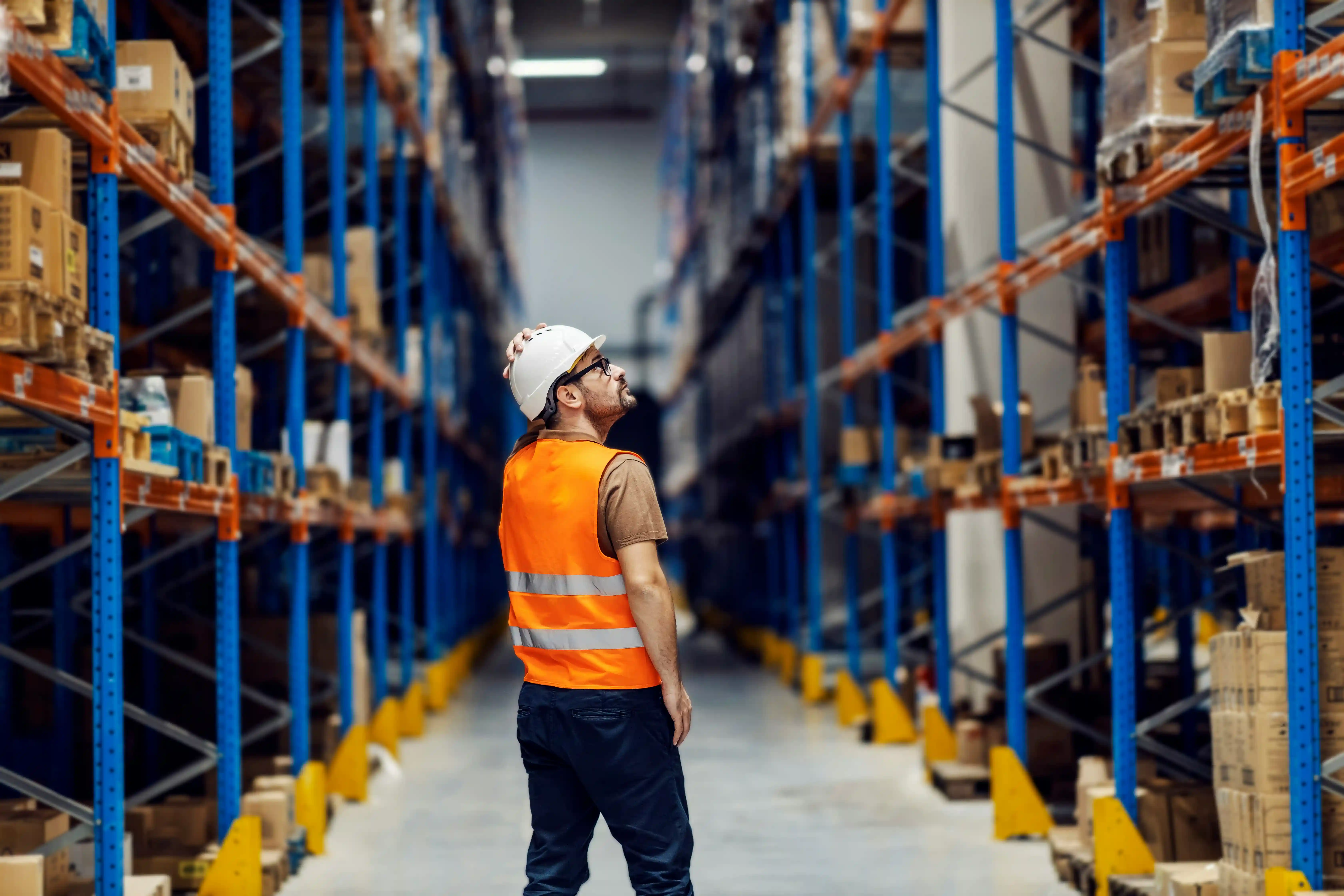 a man in a high-vis jacket and hard hat stares up a warehouse racking in a 3PL warehouse