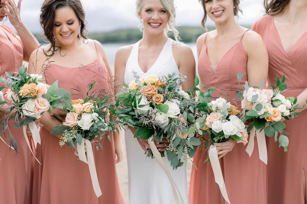 Bride in a white gown stands with bridesmaids in matching terracotta dresses, all holding bouquets of white and peach roses, greenery, and ribbons.