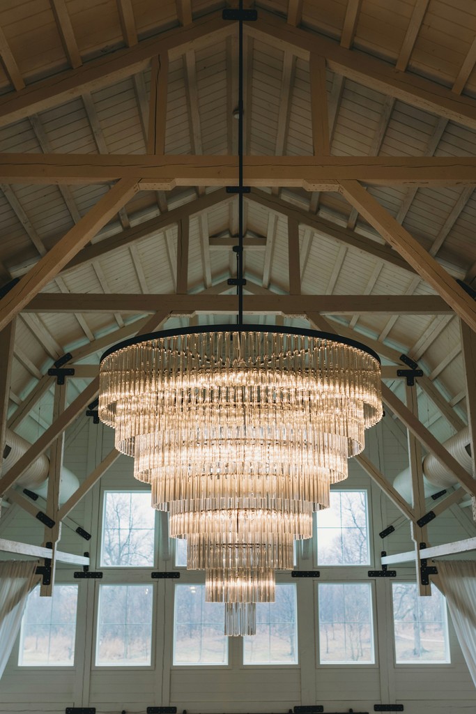 Tiered crystal chandelier hanging from exposed wooden rafters inside a bright modern barn wedding venue with tall windows