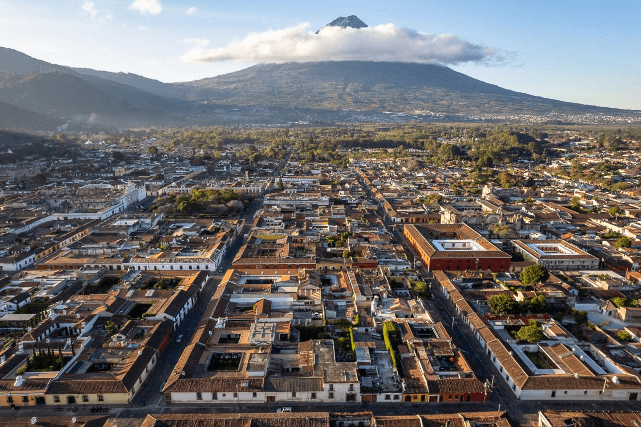 Aerial view of a colonial city with a grid-like arrangement of colorful buildings, surrounded by lush greenery and dominated by a towering volcano beneath a partly cloudy sky.
