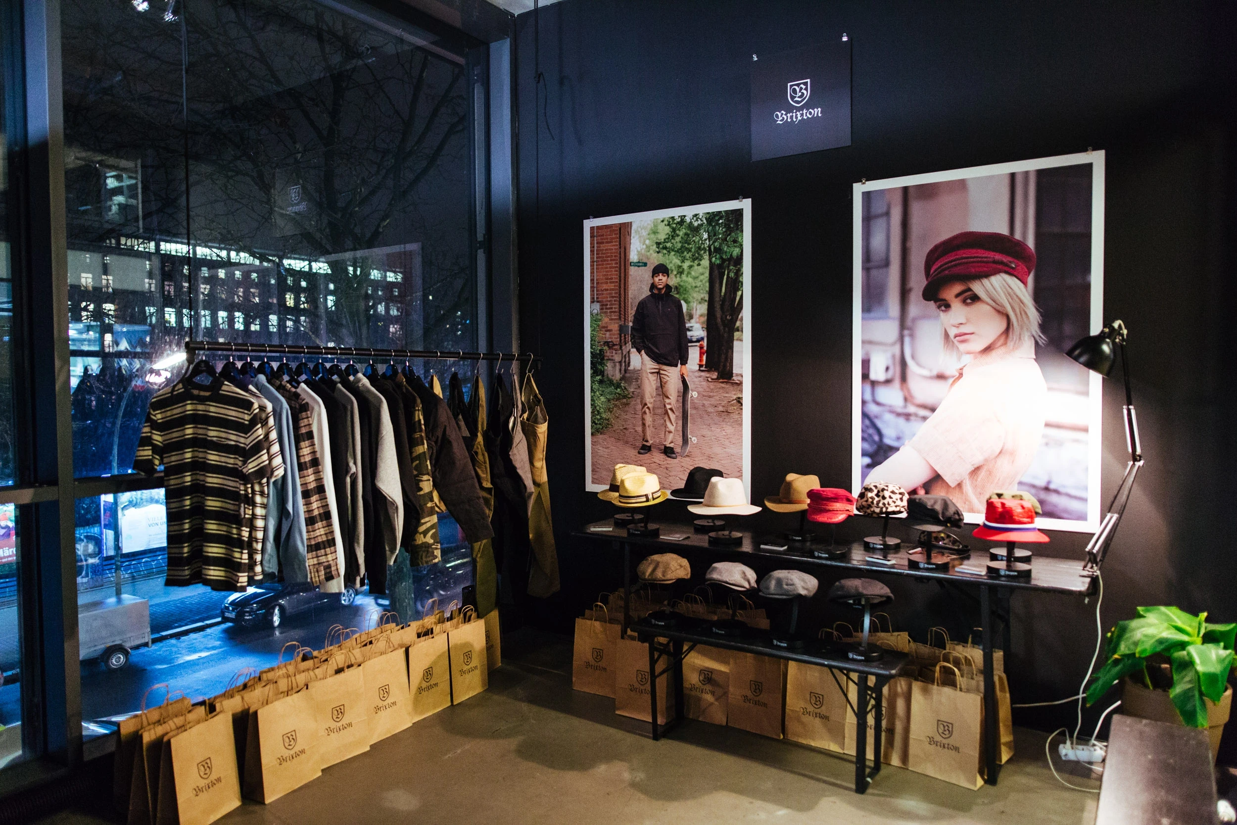 Product presentation at the BRIXTON pop-up with a clothing rack by the window, hat displays on dark benches, large campaign posters and brown paper bags.