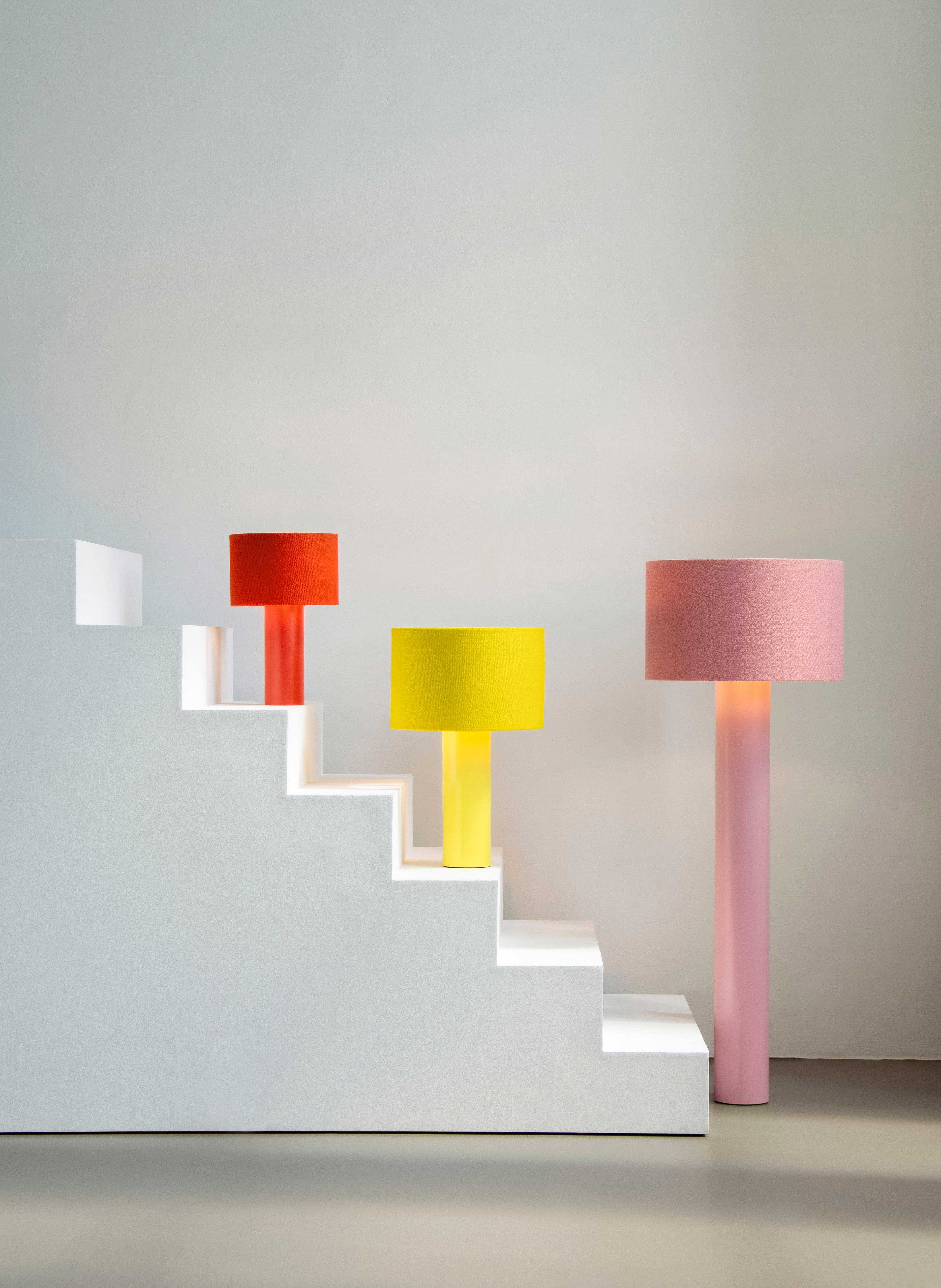 Three floor lamps with cylindrical bases and shades in red, yellow, and pink are placed on and around a white, stepped platform or staircase.