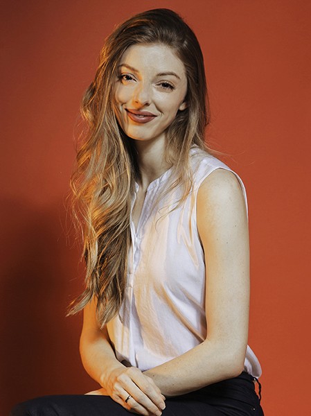 A woman with long, wavy blonde hair sits against a solid orange-brown background. She wears a sleeveless white blouse and dark-colored pants. Her right hand rests on her lap, and her left hand is placed near her left elbow. She looks directly at the camera with a slight smile.