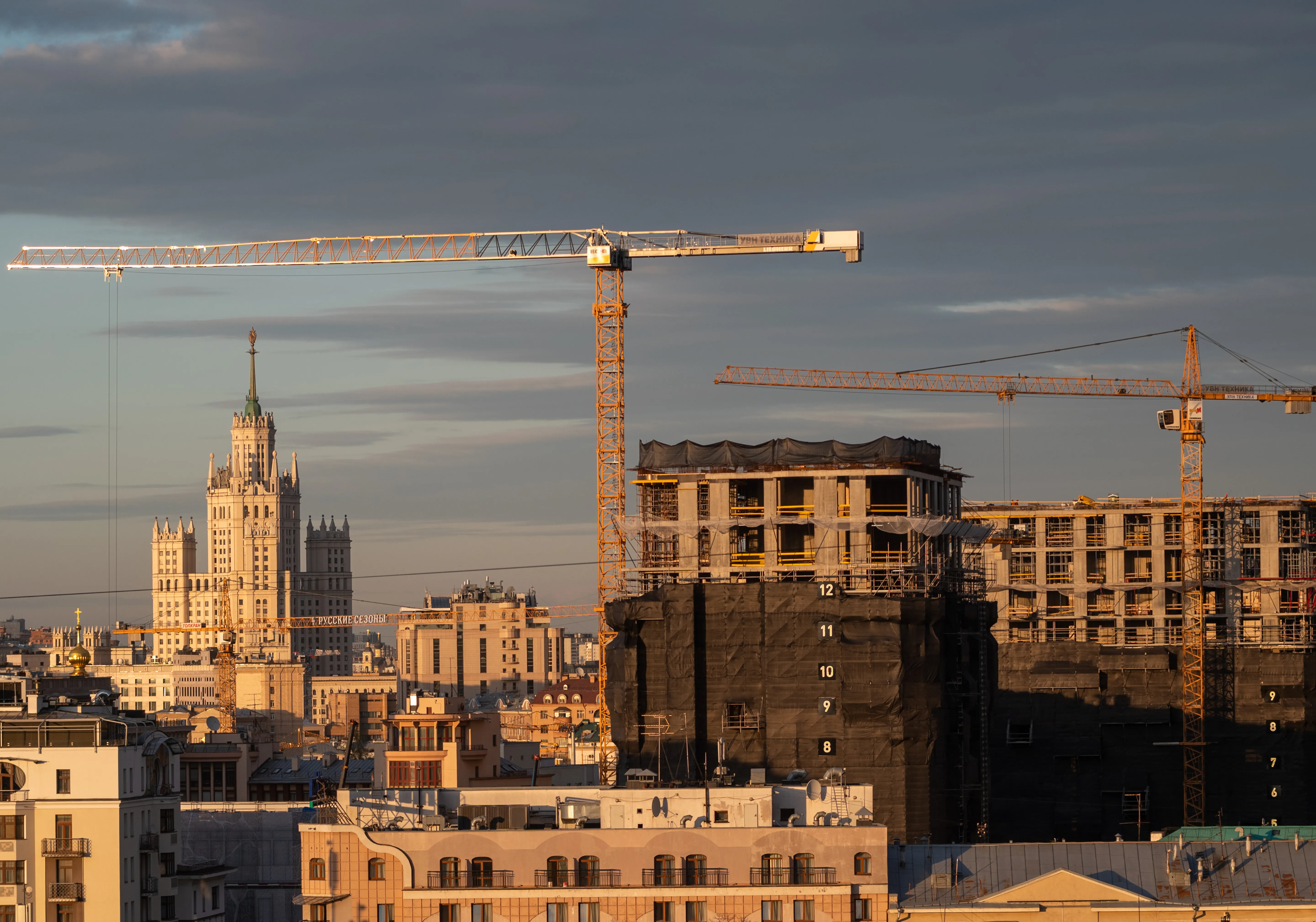 Construction cranes framing a modern building under development at dusk, with a historic skyscraper in the background.