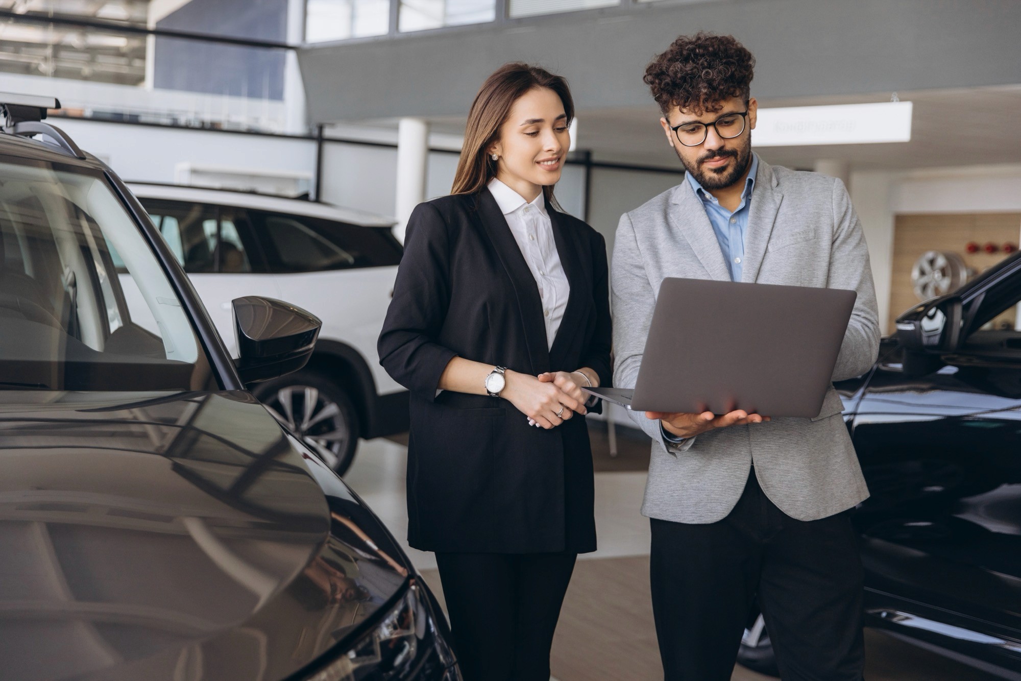 Salesman holding a laptop, assisting a customer in selecting a new car while discussing options in an elegant auto showroom