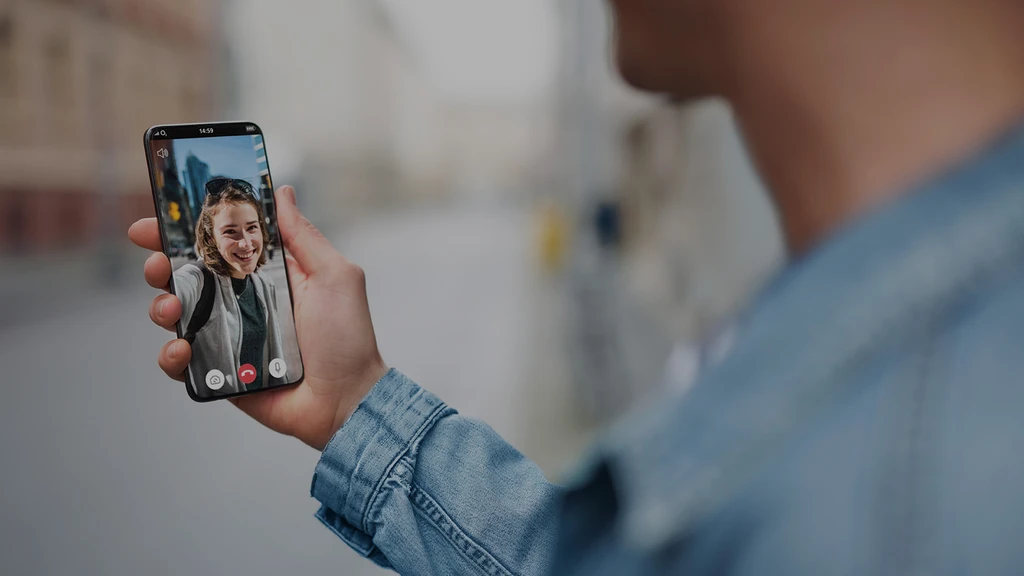 Lifestyle-Teaser für die Telefónica Case Study. Eine Person hält im Vordergrund ein Smartphone in der Hand, auf dessen Bildschirm ein lachendes Gesicht während eines Videoanrufs in einer urbanen Umgebung zu sehen ist.