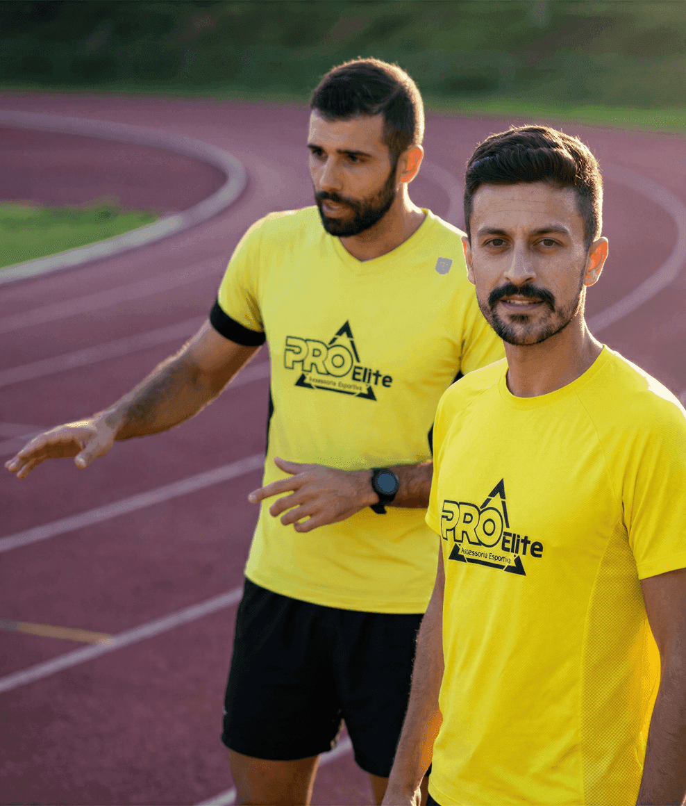 Dois treinadores de corrida de rua com camisetas amarelas da "Pro Elite" em uma pista de atletismo.