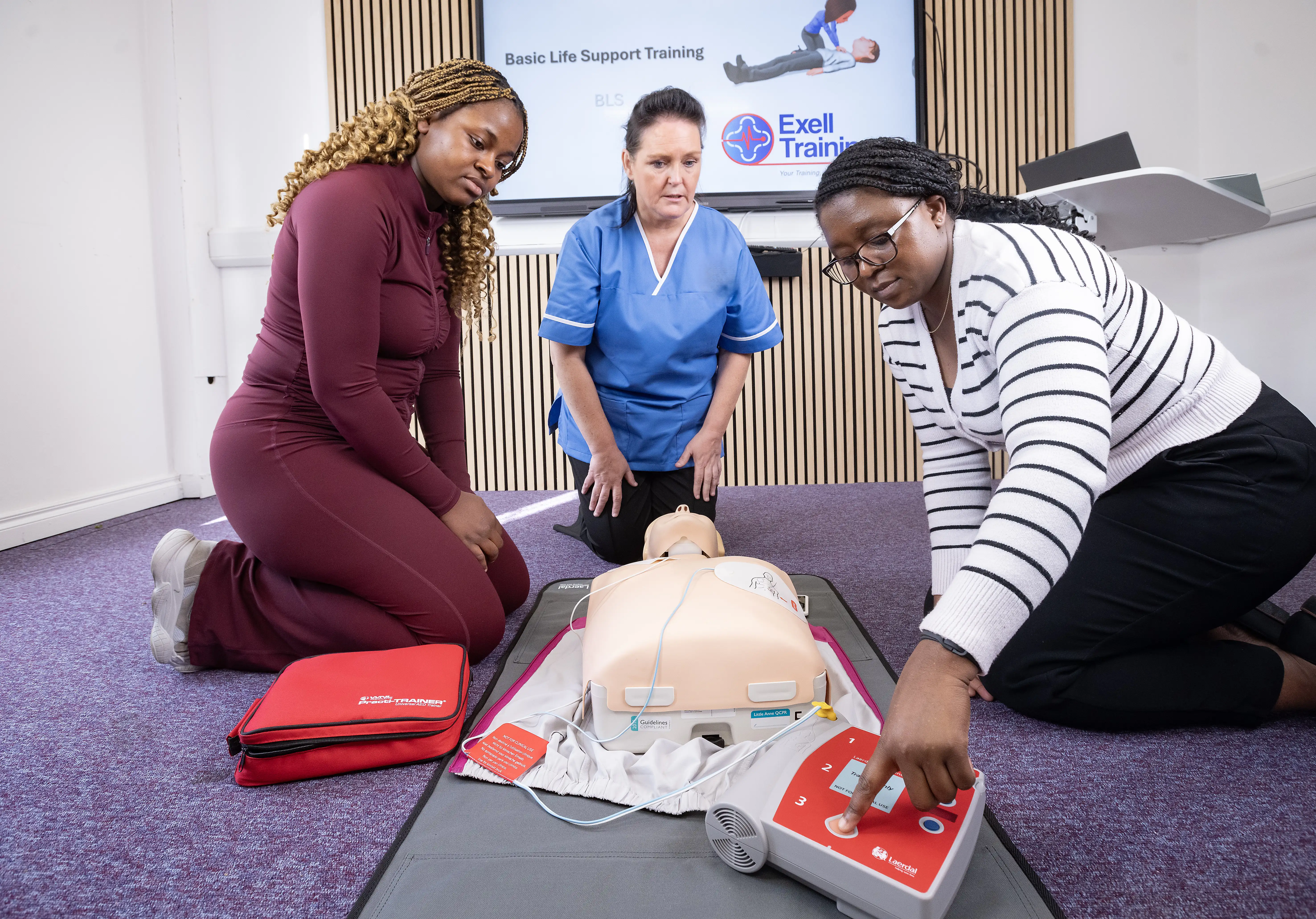 Three women practising basic life support (BLS) and AED use on a medical mannequin during a training session at Exell Training, with a defibrillator and training materials visible.