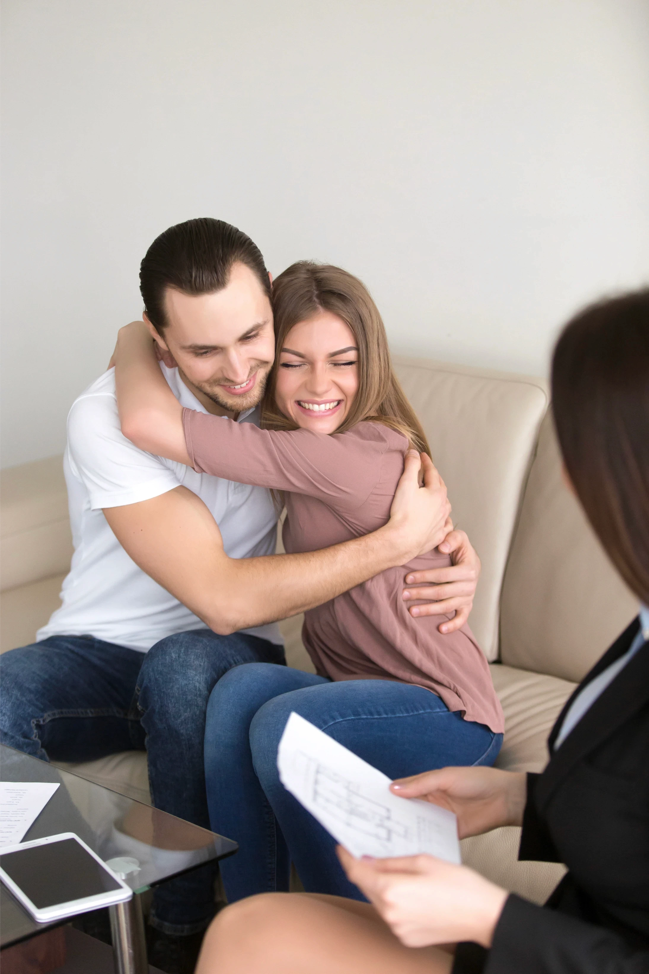 A happy couple embraces while sitting on a beige sofa, smiling. A woman in business attire sits nearby, holding documents. The mood is joyful and celebratory.
