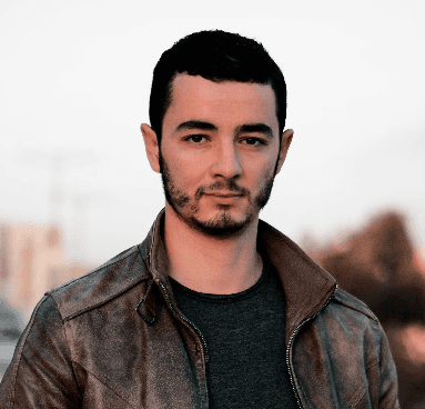 A person with curly hair and glasses smiles at the camera, seated in a cozy workspace with warm lighting.