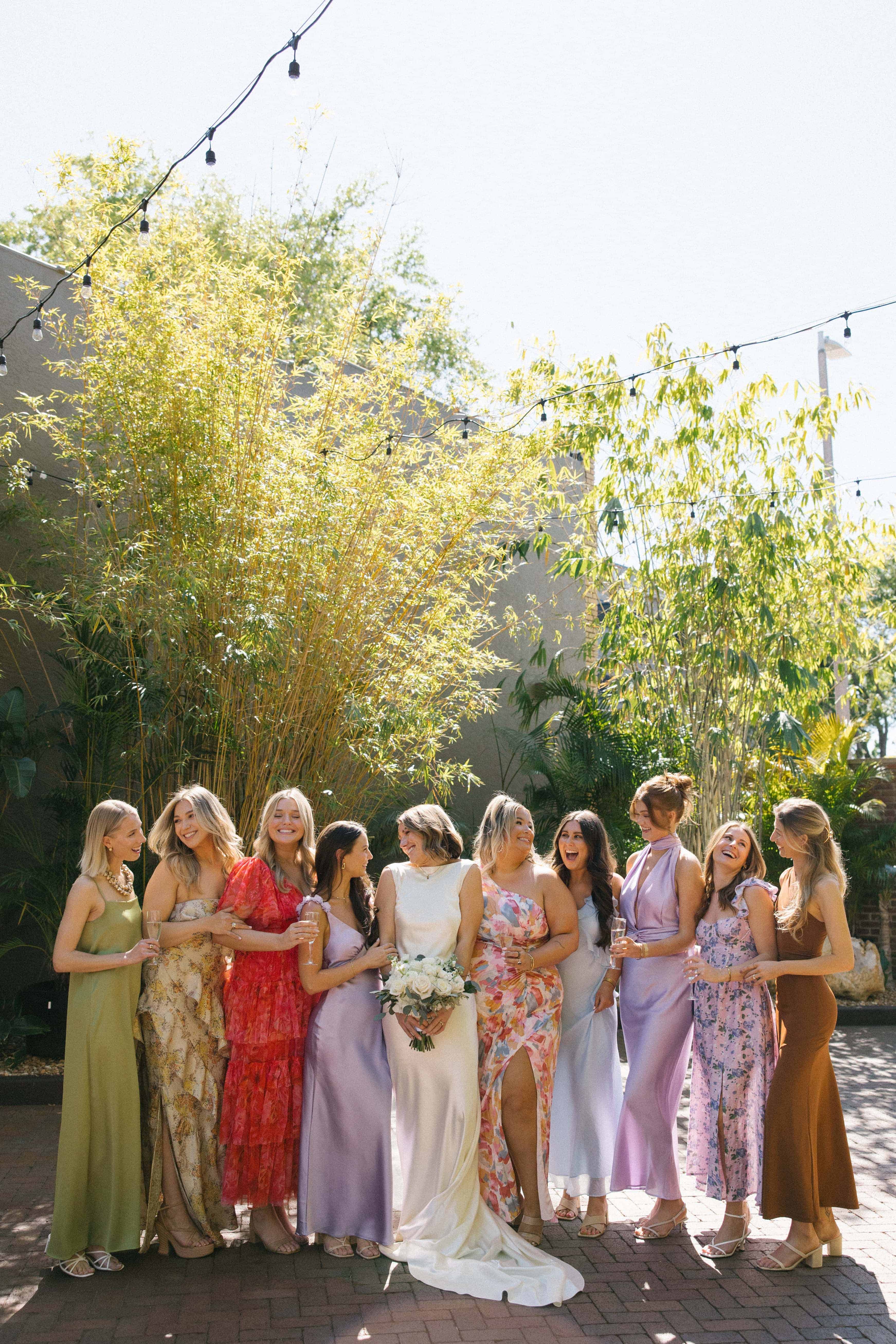 A group of women in various colored dresses are standing in a line outdoors with string lights hanging above them.