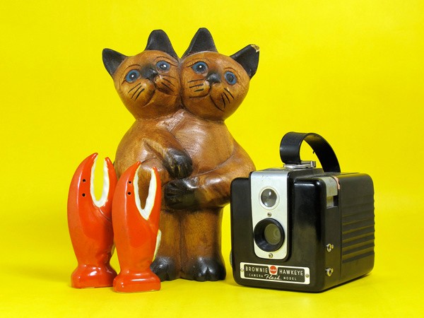 The image shows a brown cat figurine made of wood, standing upright on its hind legs and holding two claw-like objects. Next to it is a black instant camera with a silver top and label that reads "BROWNIE HAWKEYE CAMERA MODEL". The figurine and camera are set against a solid yellow background.