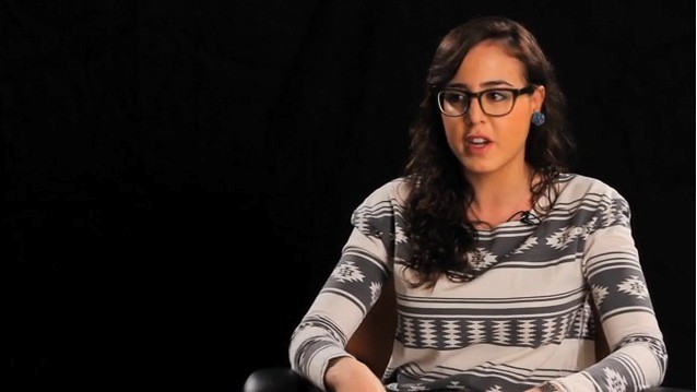 A still shot image of a woman wearing glasses, a blouse and talking.