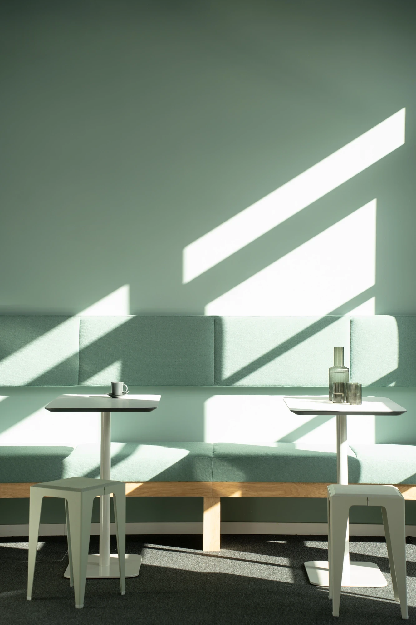 Quiet office canteen corner with mint-green banquette seating, two small white café tables and stools, lit by angled sunlight from the window.