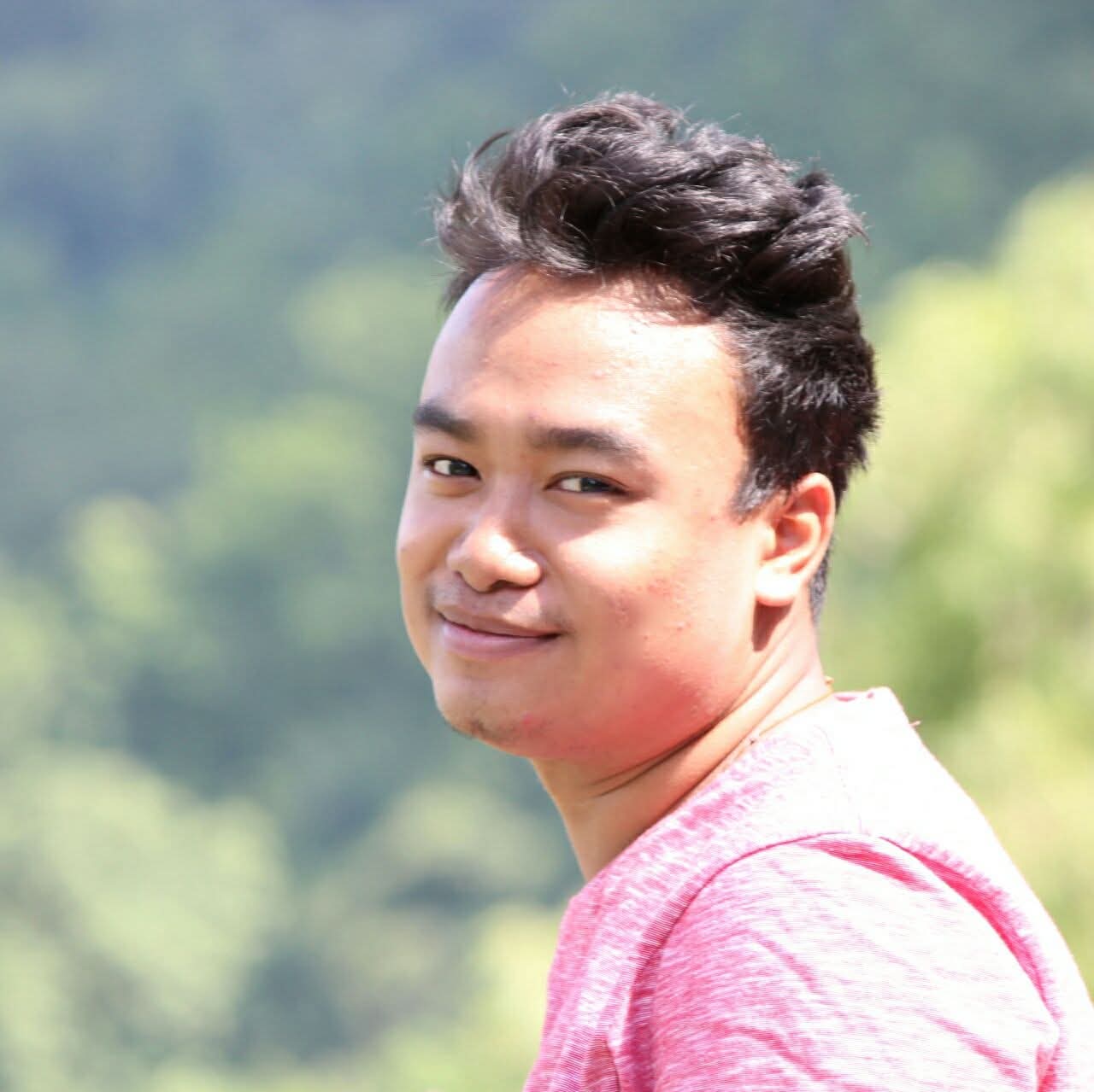 Man in a white t-shirt and cap sits at a desk under moody blue lighting. Art supplies and a framed sketch of figures under an umbrella are visible.