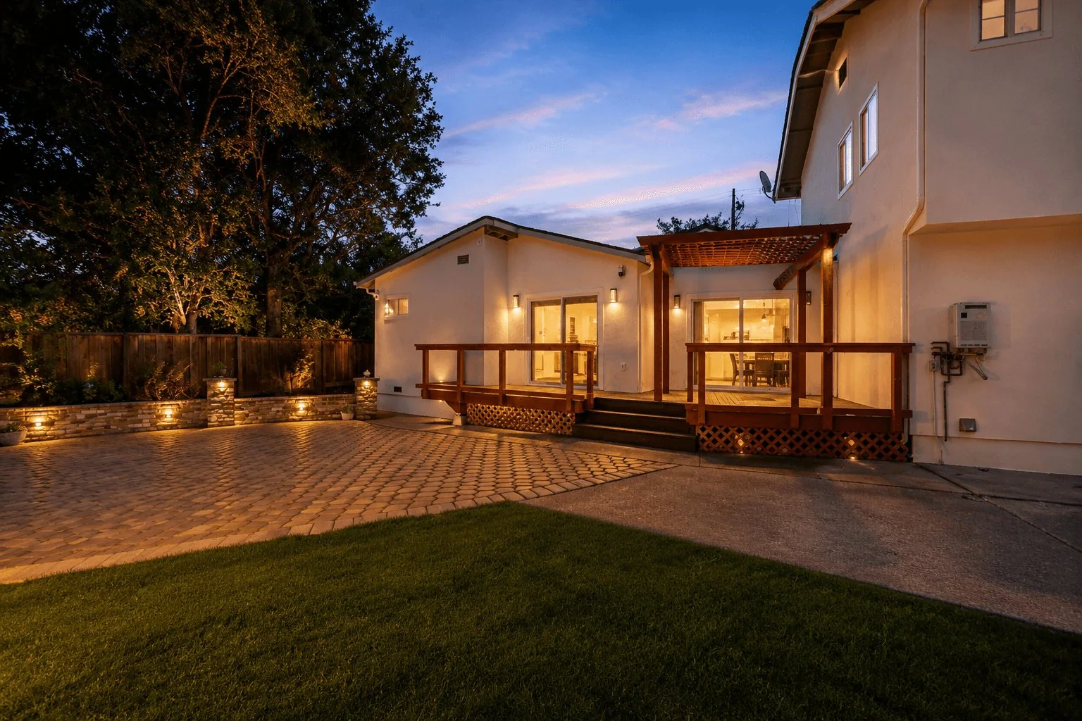 An attached ADU and the main house at dusk, with a lit wood deck and pergola