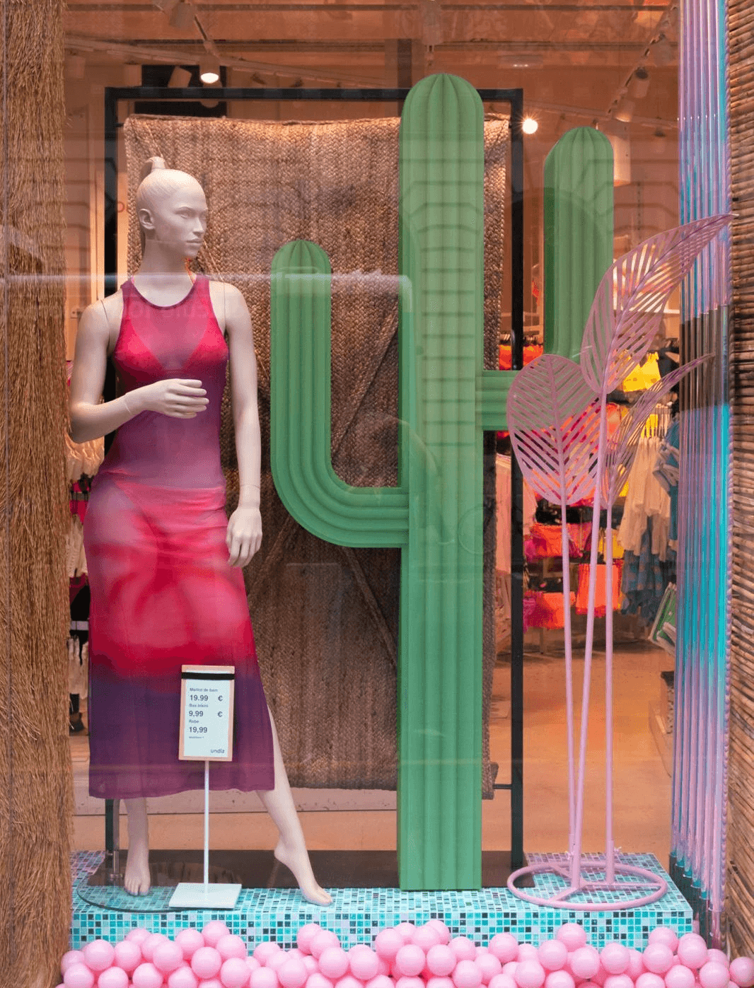 Mannequin en robe dégradée rose et violette à côté d'un cactus vert décoratif dans la vitrine Undiz.