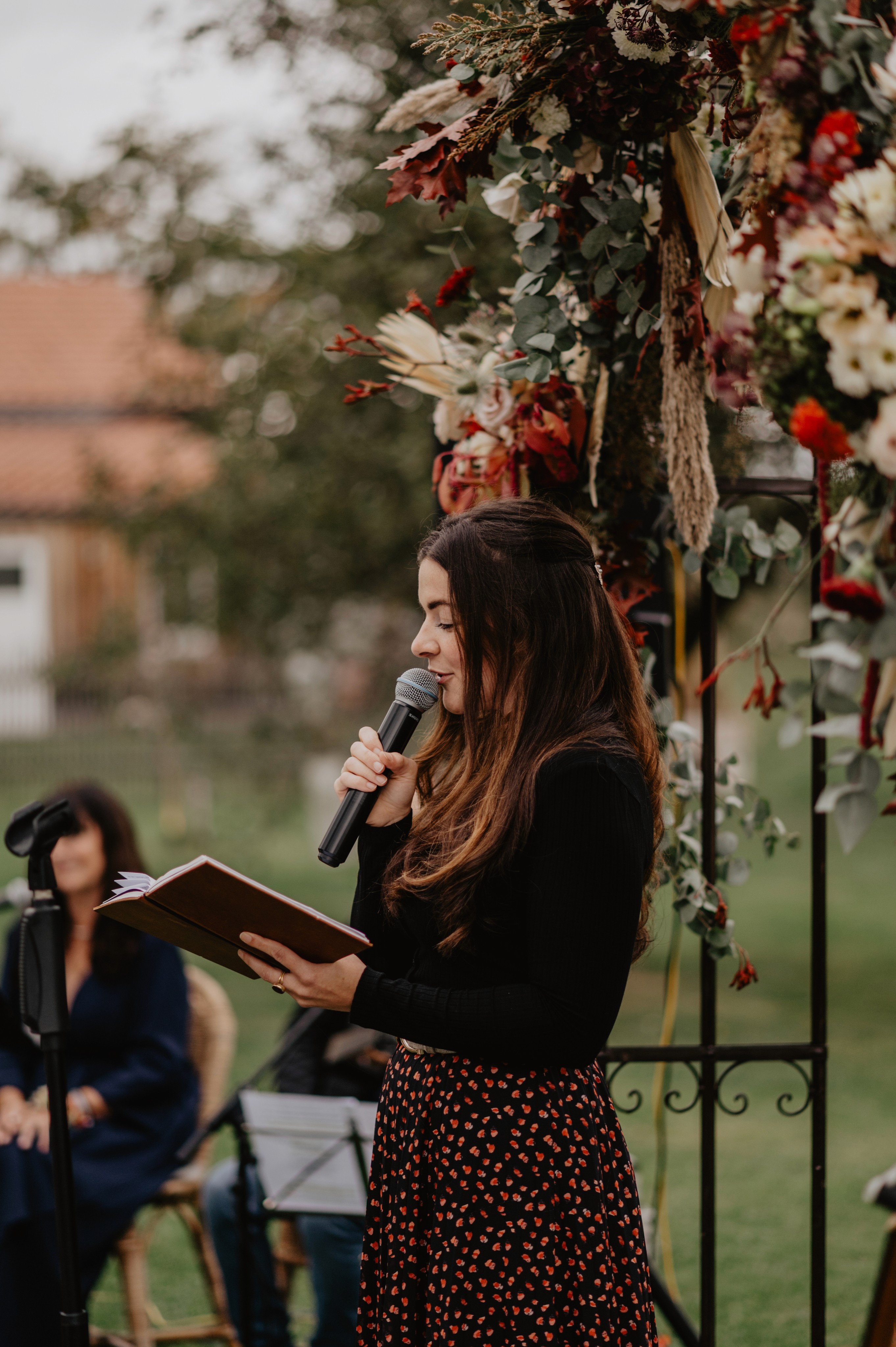 Maria Wiethaler freie Rednerin und Sängerin für Hochzeiten, Taufen, Trauerfeiern und Events aus München und Landshuter Umgebung. Eine Frau mit langen, welligen braunen Haaren und einem schwarzen Outfit hält ein Mikrofon und ein aufgeschlagenes Notizbuch, während sie eine Rede hält. Sie steht unter einem floral geschmückten Bogen mit roten, weißen und grünen Blumen sowie getrockneten Gräsern. Im Hintergrund sitzt eine weitere Frau mit dunklen Haaren und einem schwarzen Outfit, während eine ländliche Kulisse mit einem Gebäude und Bäumen zu sehen ist. Die Szene wirkt feierlich und emotional.