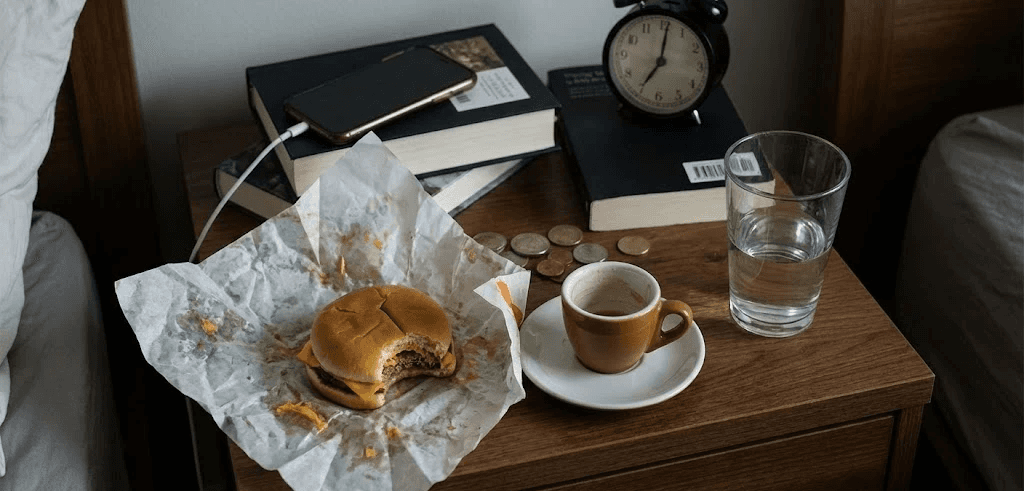 Chaotic bedside table: half-eaten burger wrapper, empty espresso cup, glass of water.